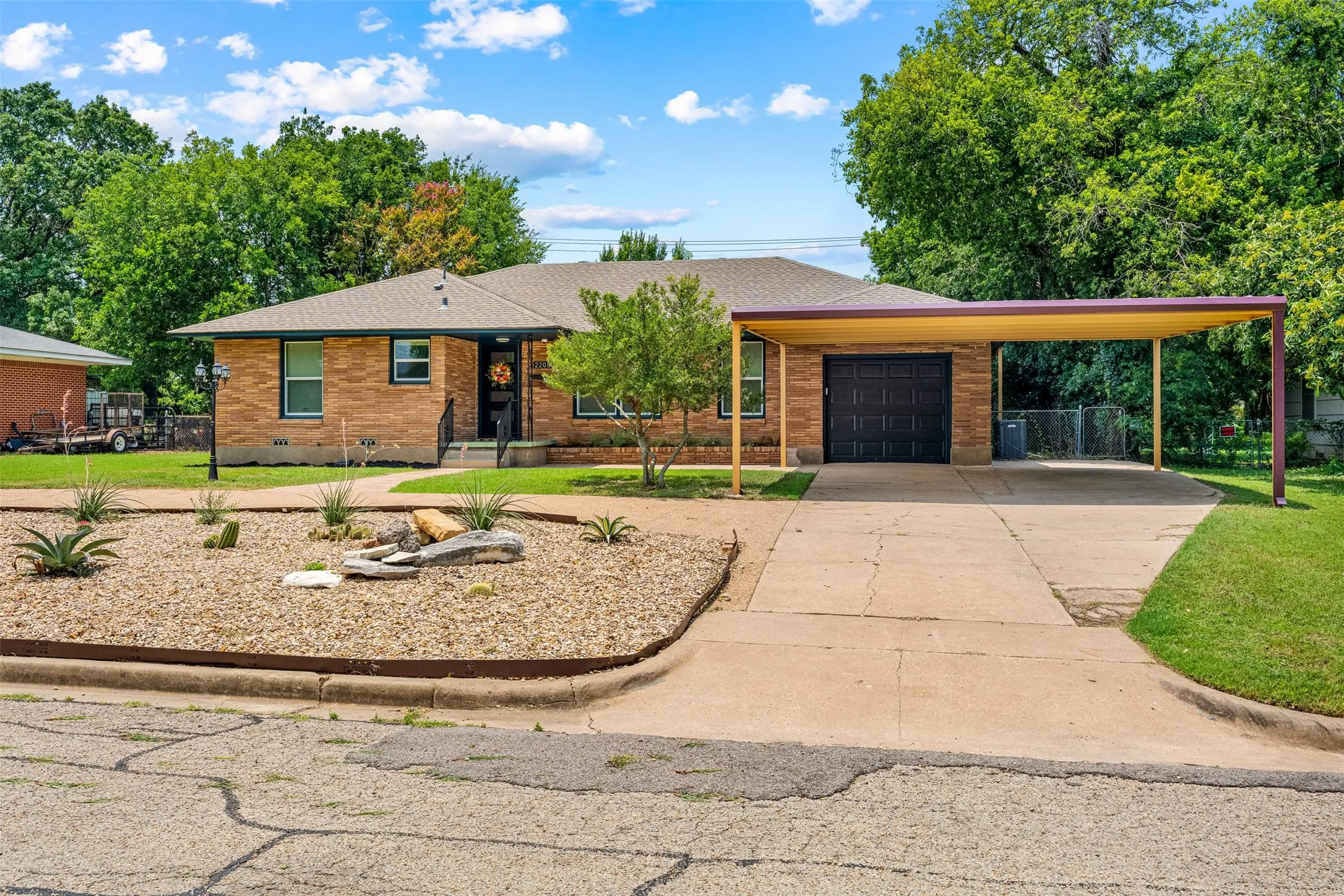 Brick exterior residence featuring a carport, an attached garage with a dark panel door, a concrete driveway, and a front yard with a xeriscape landscaping bed