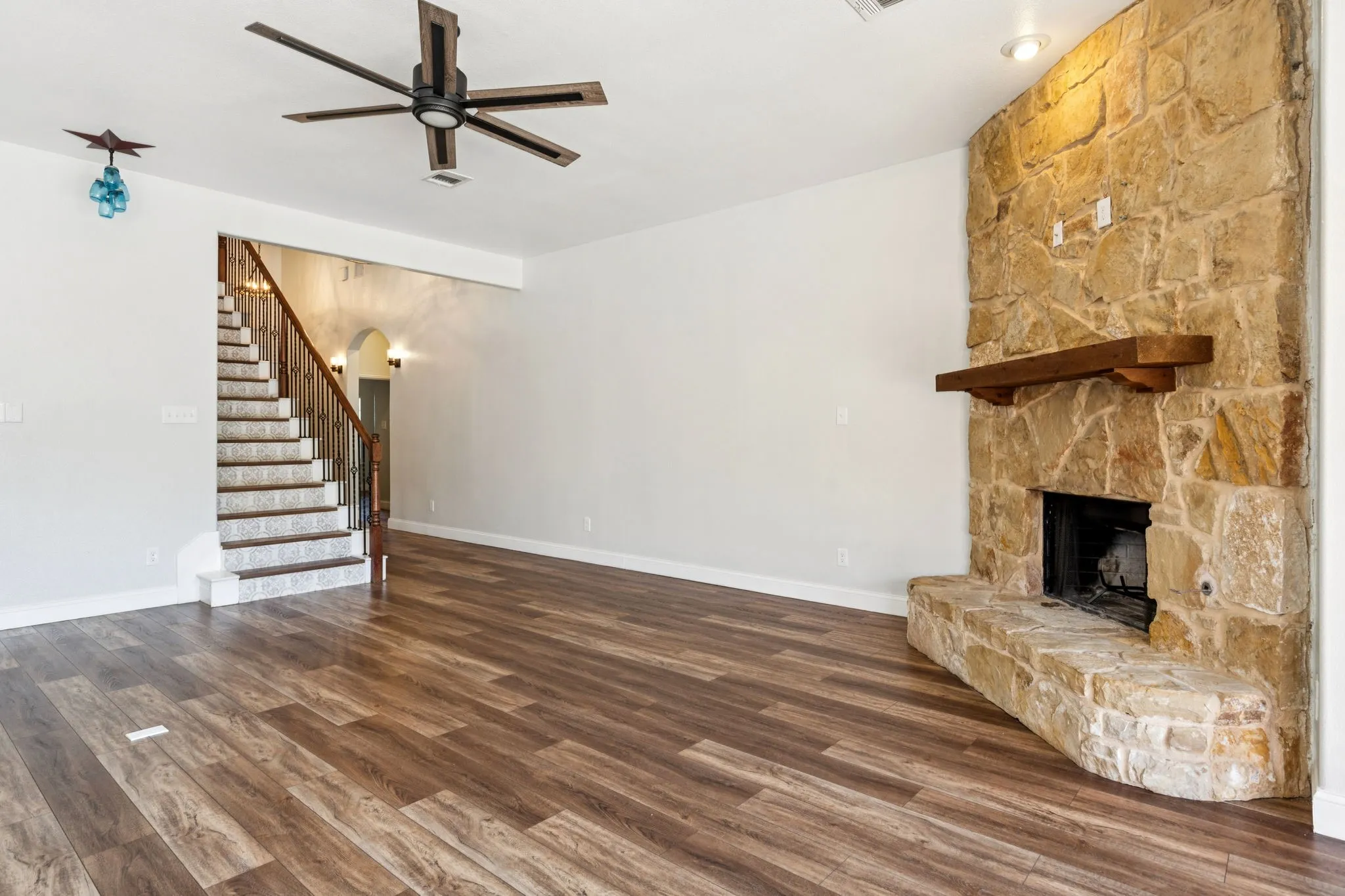 Unfurnished living room featuring a stone fireplace, dark wood finished floors, ceiling fan, stairway, and arched walkways