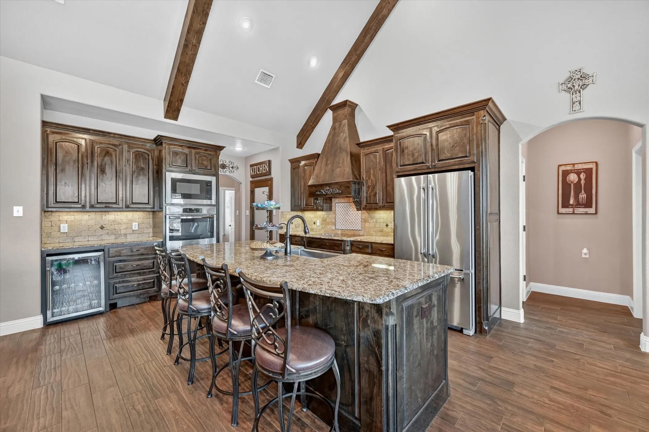 Spacious kitchen featuring an expansive center island with seating, a built-in beverage cooler, stainless steel appliances, a custom range hood, and vaulted ceilings with exposed wood beams