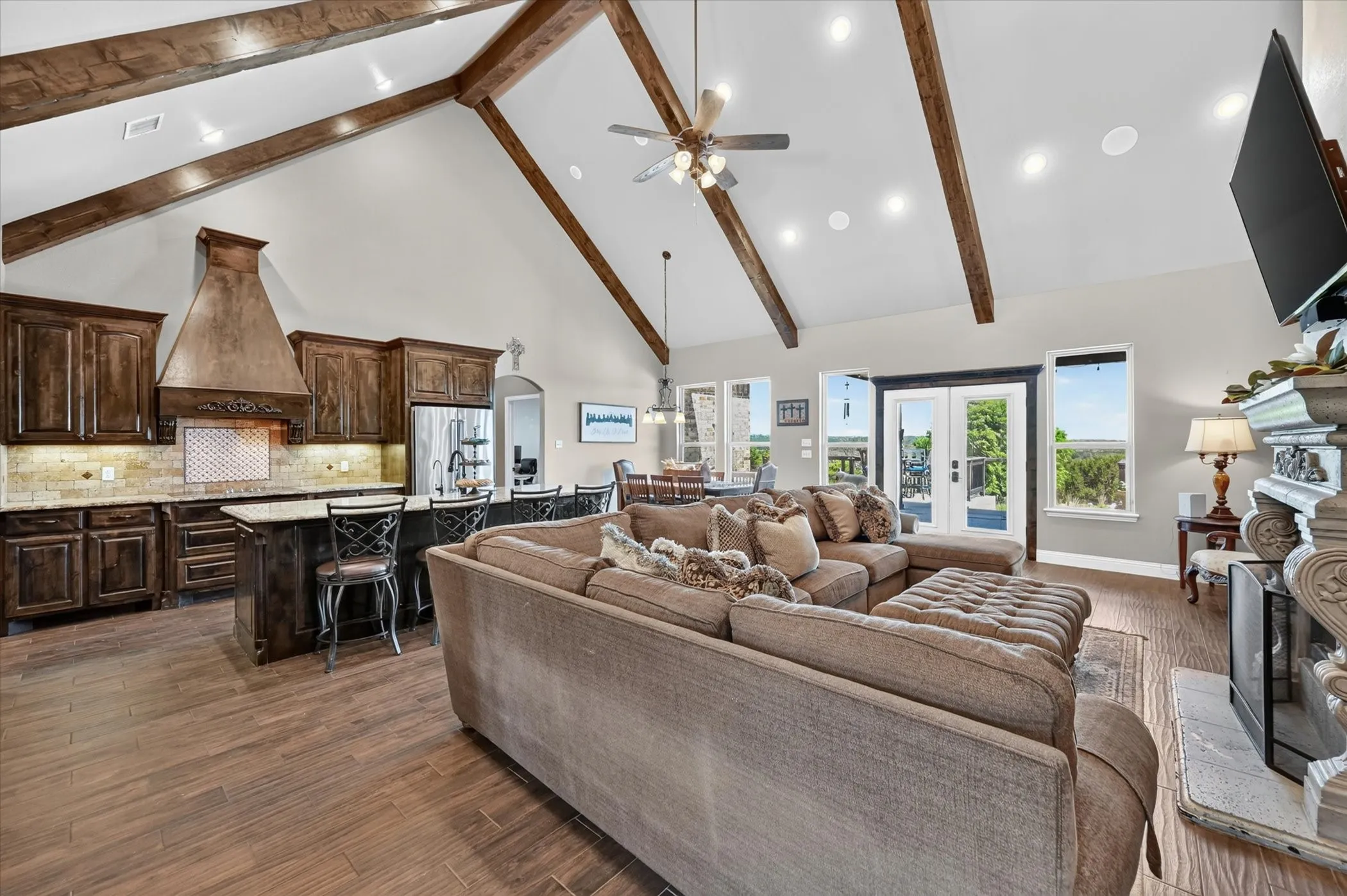 Vaulted ceiling with exposed wood beams, wood-finish flooring, built-in fireplace, kitchen island with seating, and double glass doors opening to the exterior