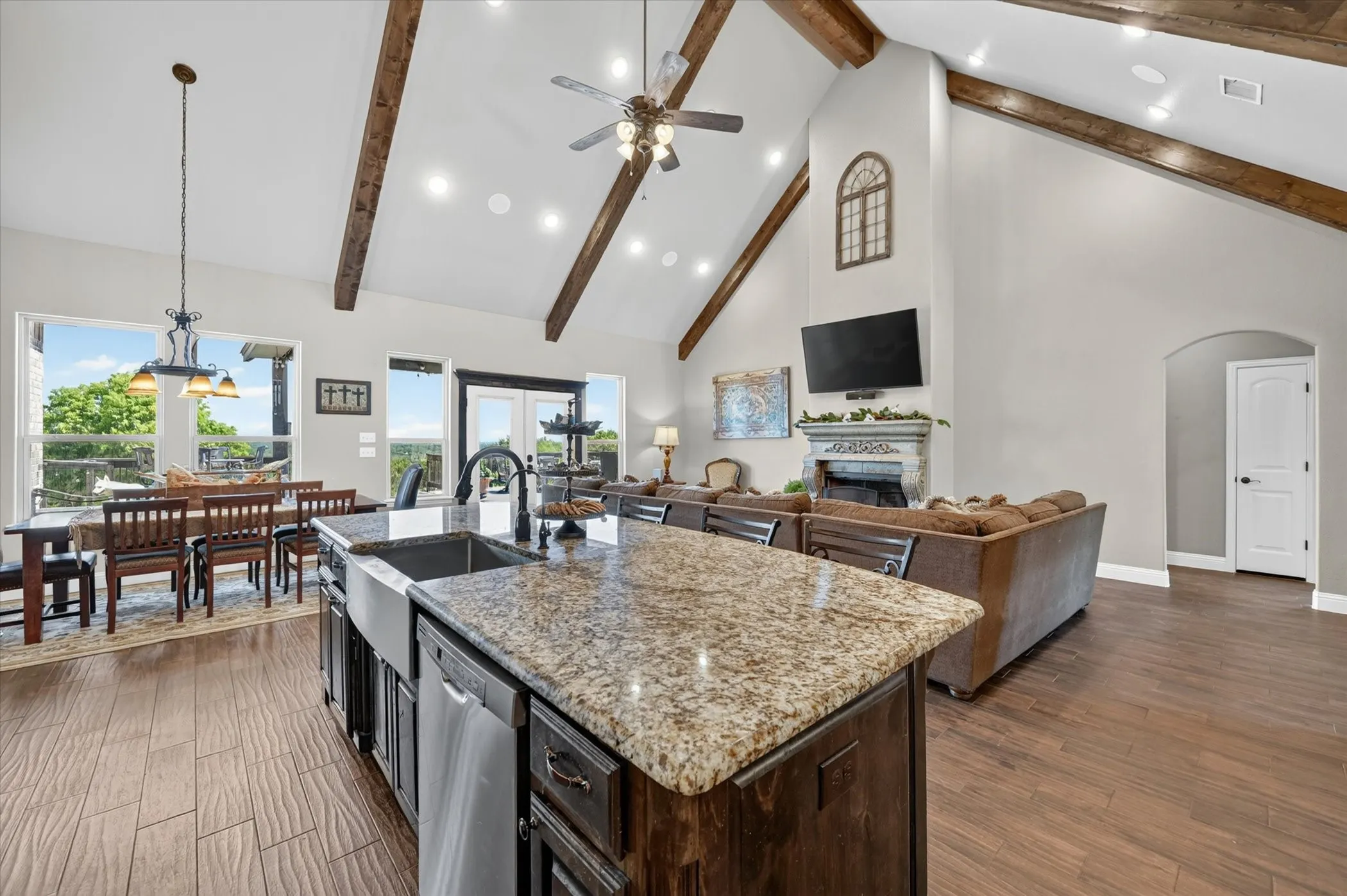 Spacious open-concept interior featuring vaulted ceilings with exposed wood beams, an expansive kitchen island with a farmhouse sink and granite countertop, stainless steel appliances, and wood-finish flooring