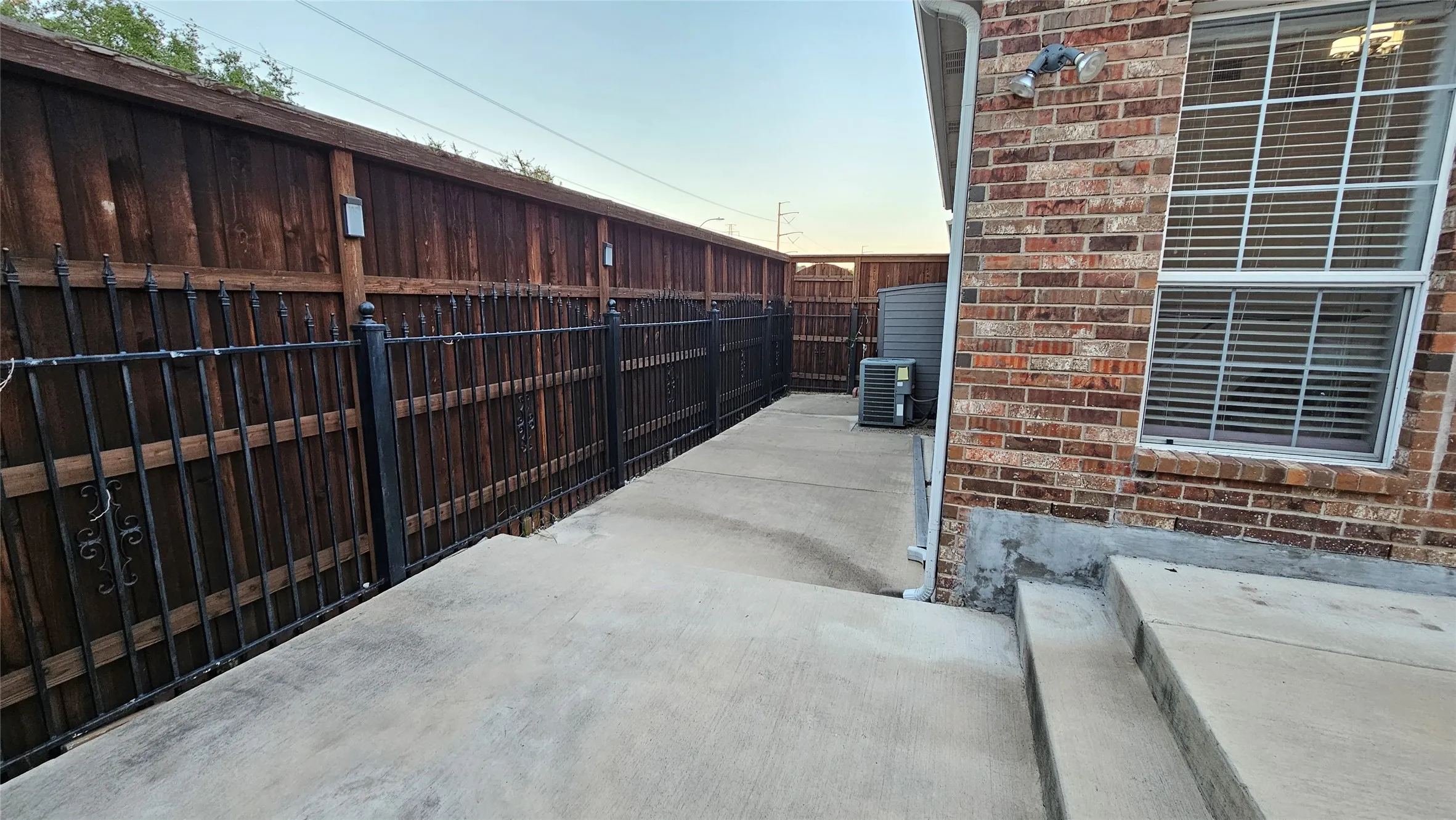 Concrete patio area featuring a brick exterior wall, a window with horizontal blinds, and a durable wood privacy fence with decorative wrought iron accents