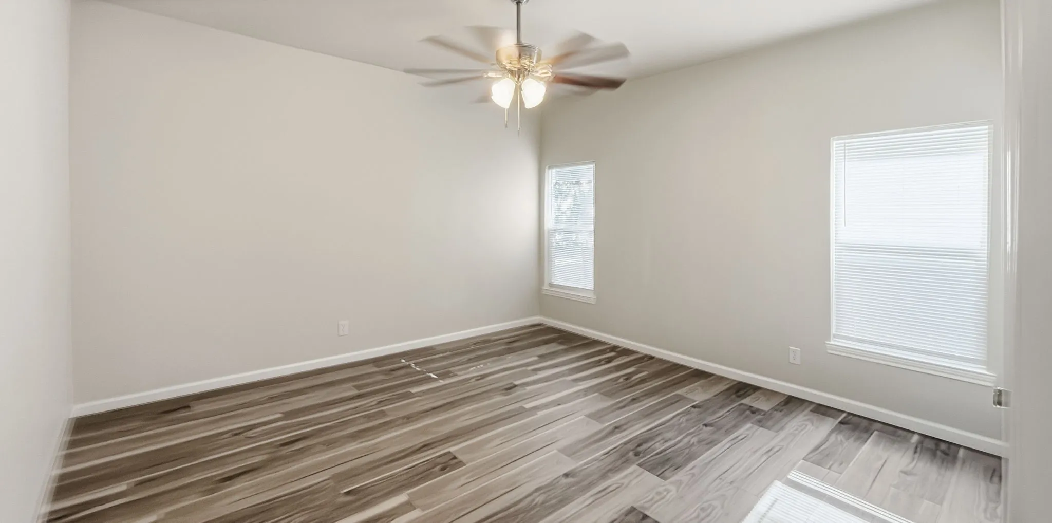 Spacious room featuring wood-finish flooring, two windows with blinds, a ceiling fan with integrated lighting, and neutral wall paint