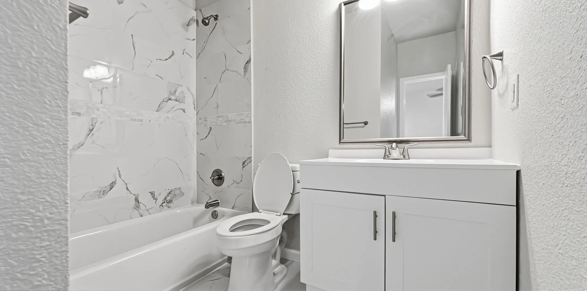 Bathroom featuring a white vanity with a single sink, an integrated bathtub and shower with white marbled tile surround, and a chrome-framed mirror
