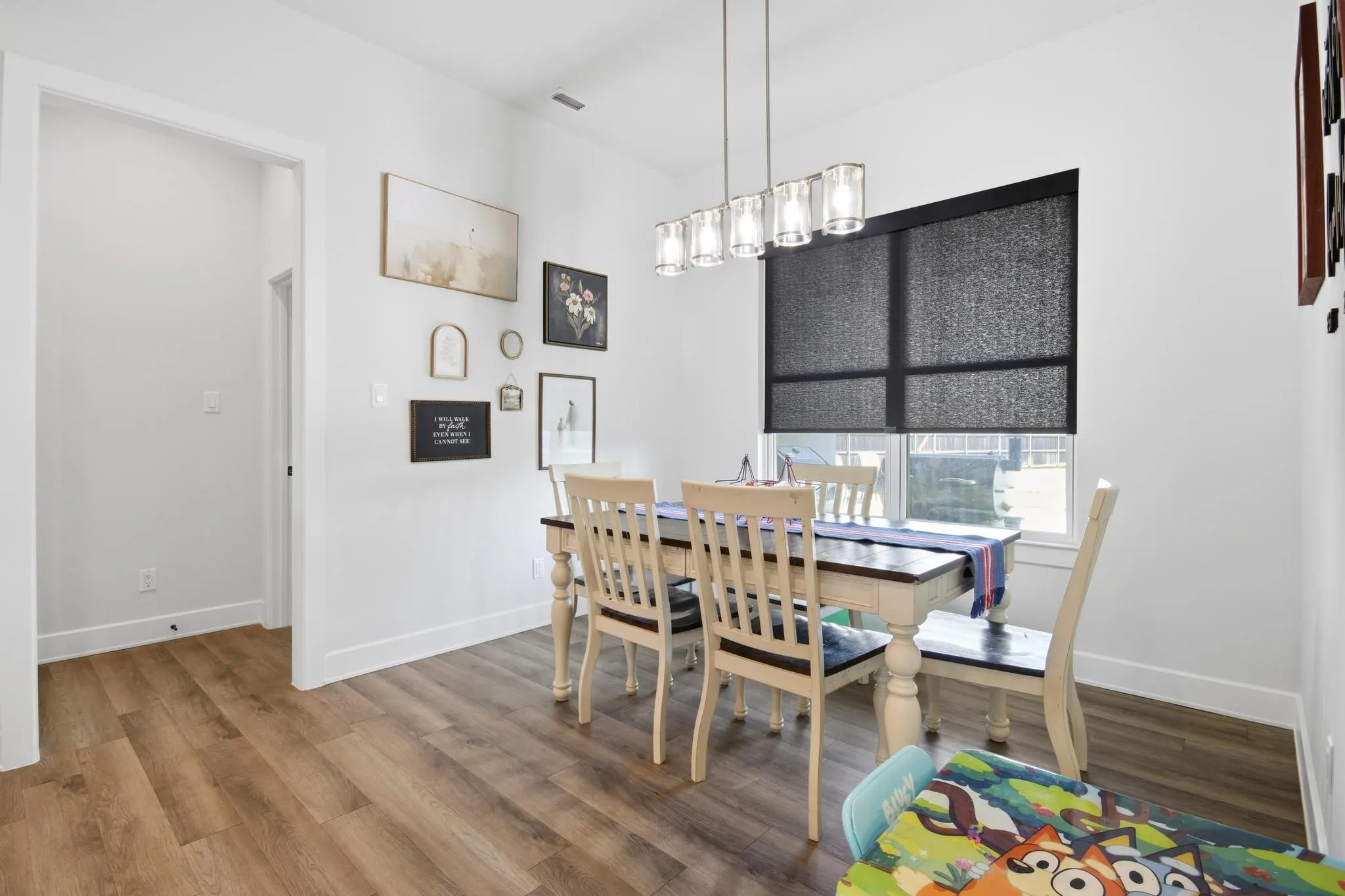 Dining area featuring wood-finish flooring, a contemporary linear chandelier, white baseboards, and a window with a dark roller shade