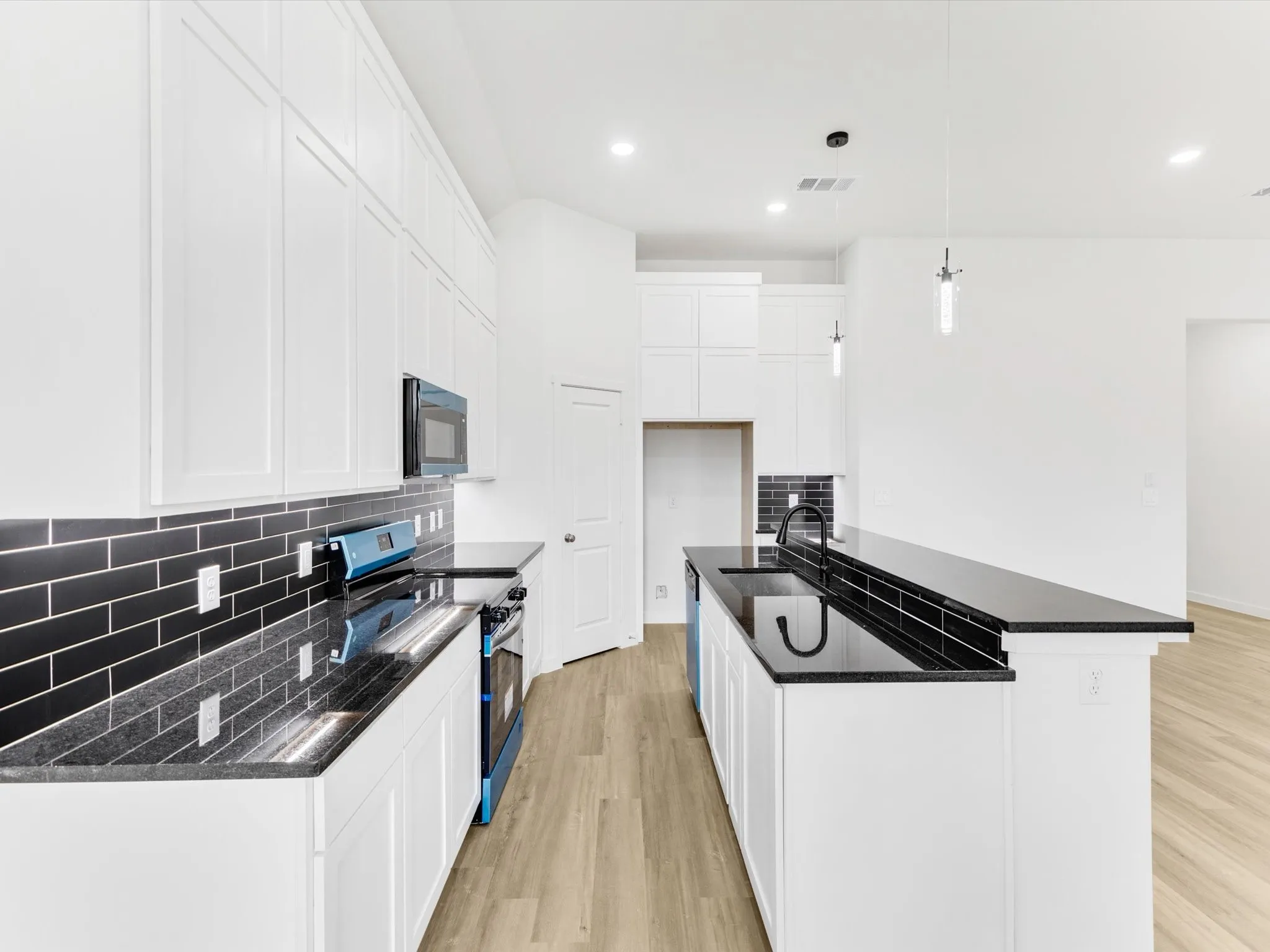 Kitchen featuring white shaker-style cabinetry, black subway tile backsplash, black countertops, a kitchen island with sink, and wood-finish flooring