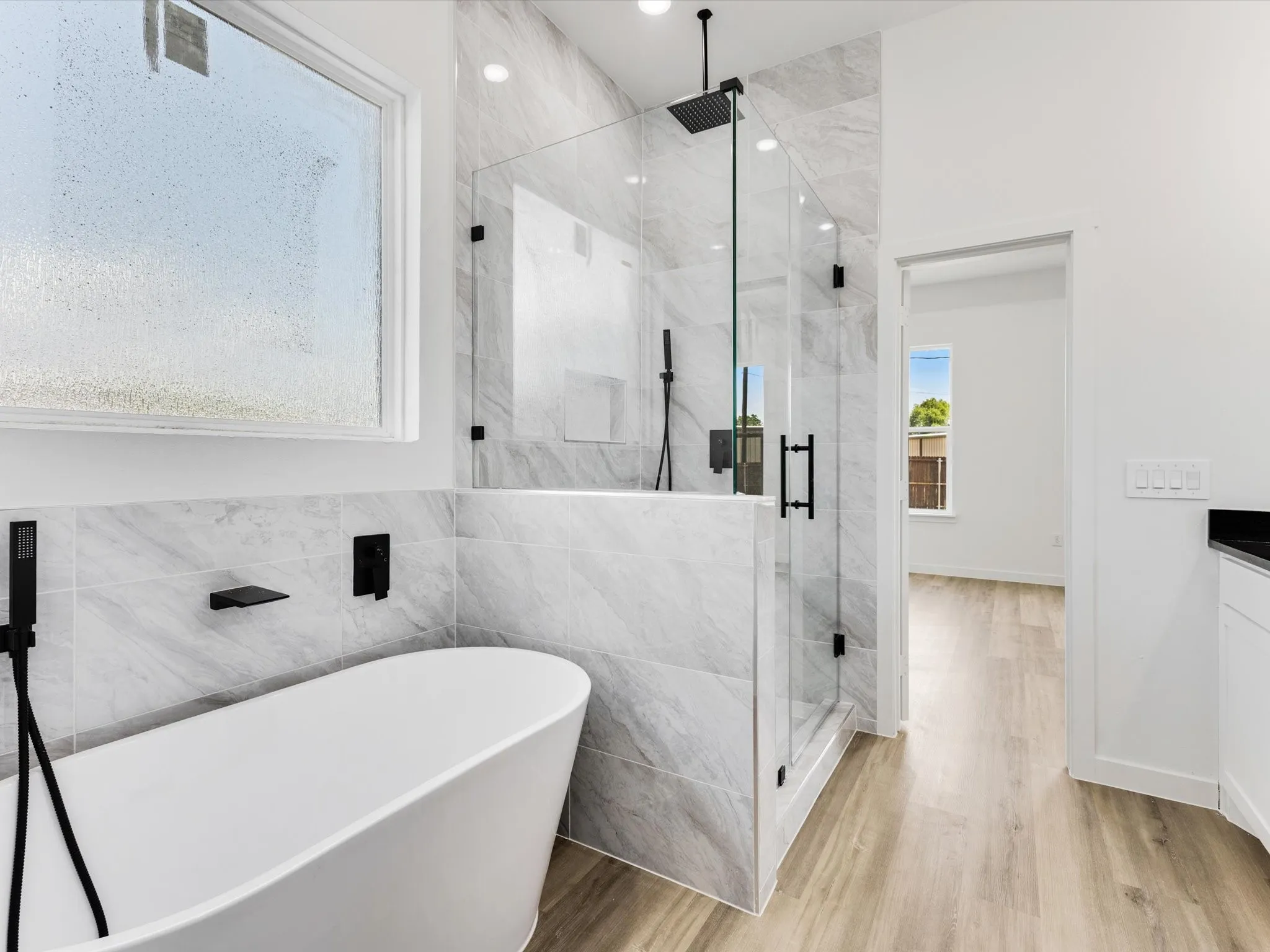 Freestanding soaking tub with matte black fixtures, glass-enclosed shower with a rainfall showerhead, large format wall tile, wood-finish flooring, and a window with privacy glass