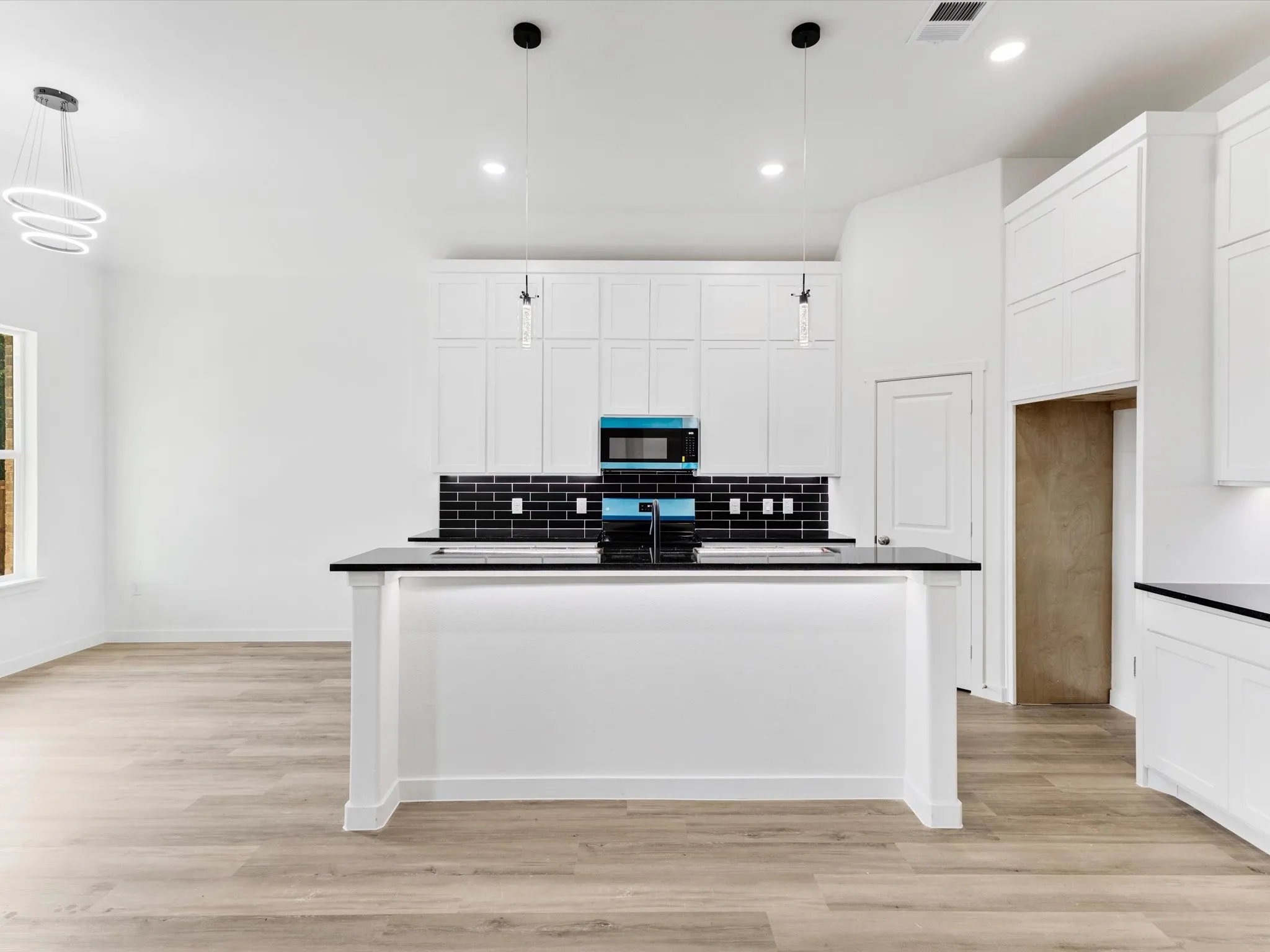 Modern kitchen featuring a large waterfall-edge island with black countertops, white shaker cabinetry throughout, stainless steel appliances, subway tile backsplash, pendant lighting, and wide-plank wood-finish flooring that extends into the adjoining living space