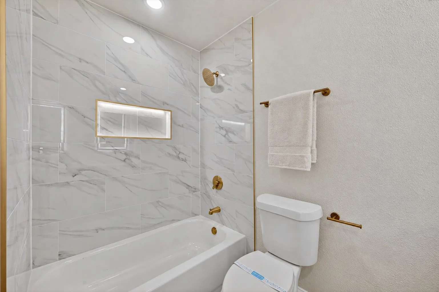 Bathtub with a white marble-look tiled surround, gold-finish fixtures, and an illuminated recessed niche