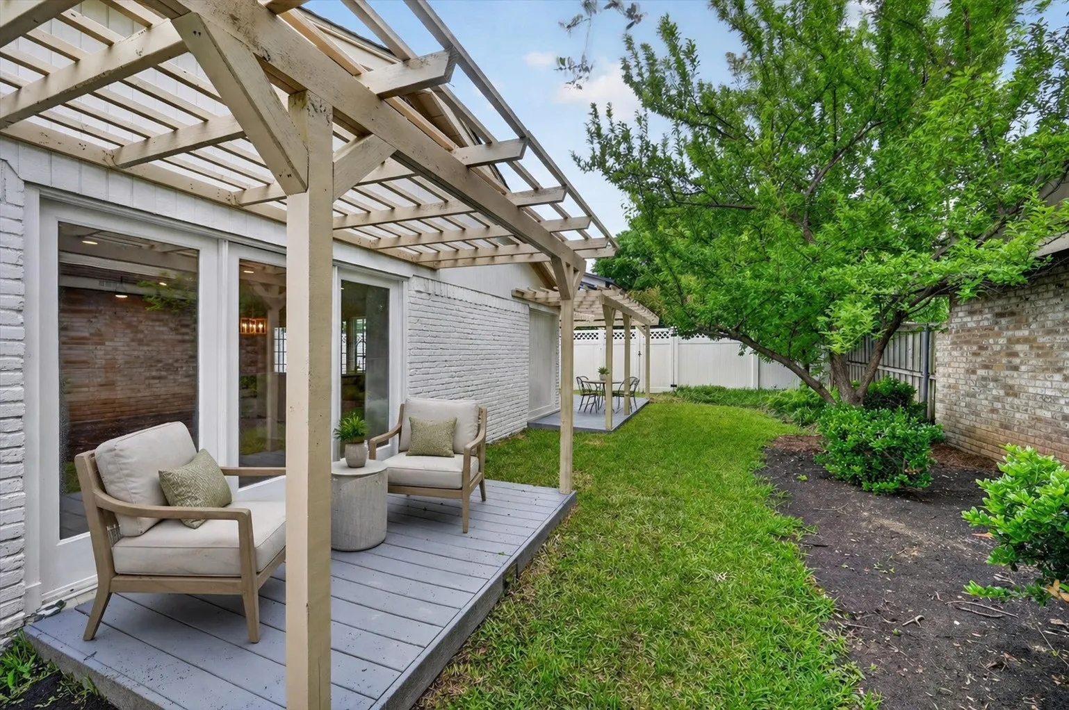 Manicured backyard featuring a pergola-covered patio with white brick exterior, sliding glass doors, and mature greenery