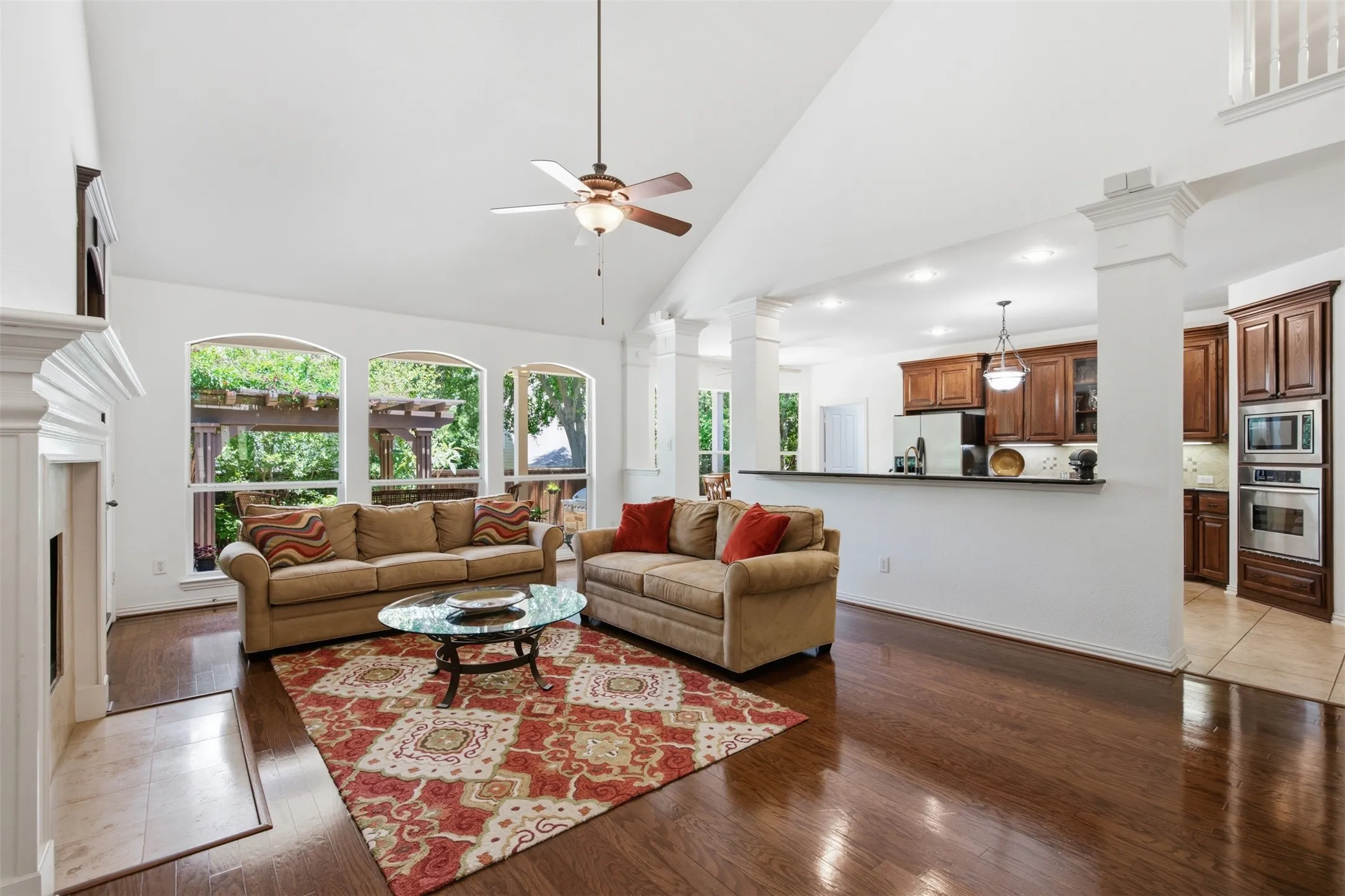 Living area featuring high ceilings, wood-finish flooring, a grand staircase with white balusters and wood handrails, a loft overlook, and a partial view of the kitchen with dark wood cabinetry and stainless steel appliances