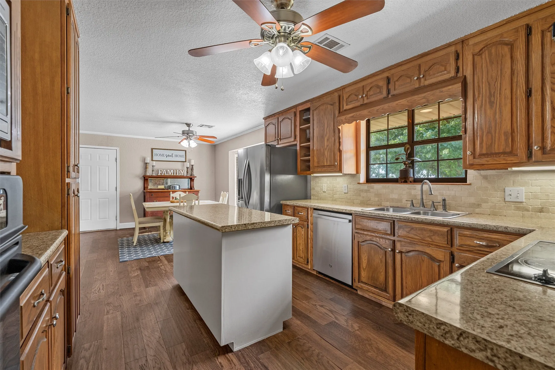 Kitchen featuring wood-finish cabinetry, light-toned countertops, a stainless steel sink, and a tile​​‌​​​​‌​​‌‌​‌‌​​​‌‌​‌​‌​‌​​​‌​​ backsplash