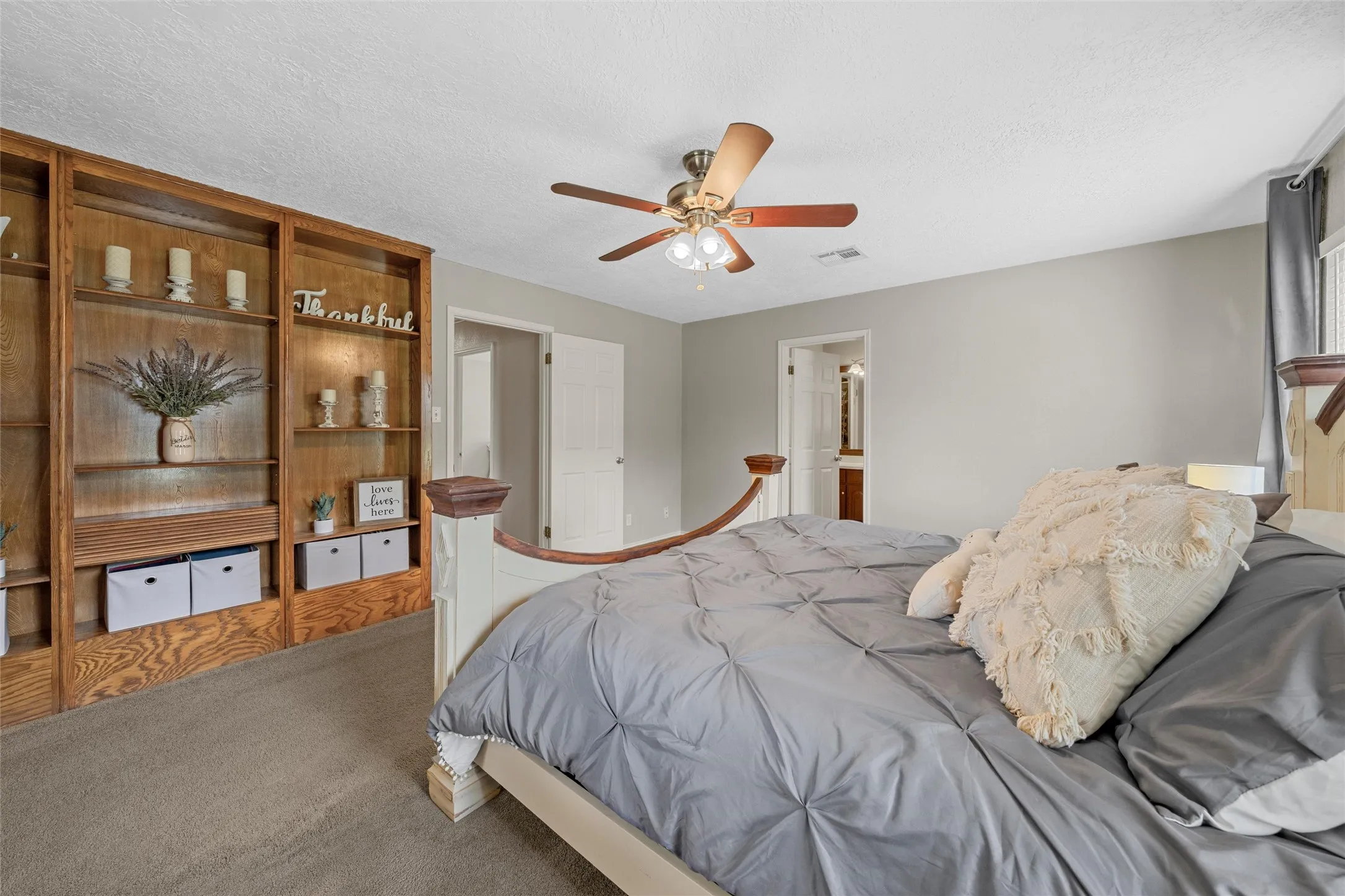 Built-in wood shelving system, ceiling fan with light fixture, light gray wall paint, carpeted flooring, and a window with dark curtain rods