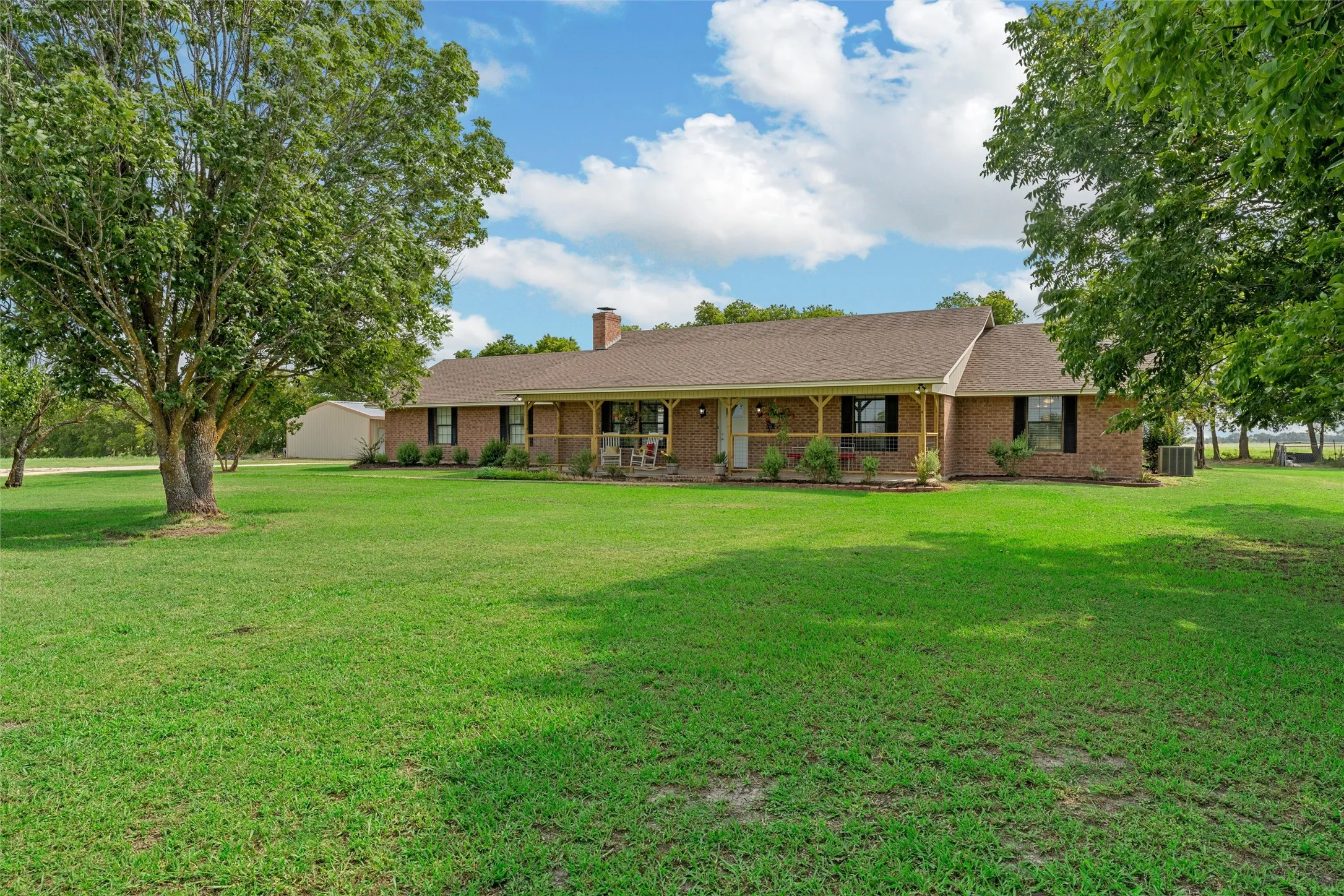 Expansive green lawn surrounds a brick residence featuring a covered front porch with wood posts and railings, dark window shutters, and a prominent brick chimney