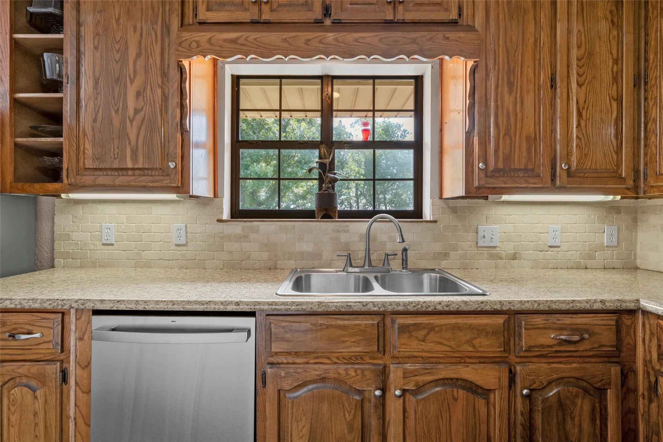 Kitchen workspace featuring a double basin stainless steel sink, a stainless steel dishwasher, wood-finish cabinetry, light-toned countertop, and a tiled backsplash