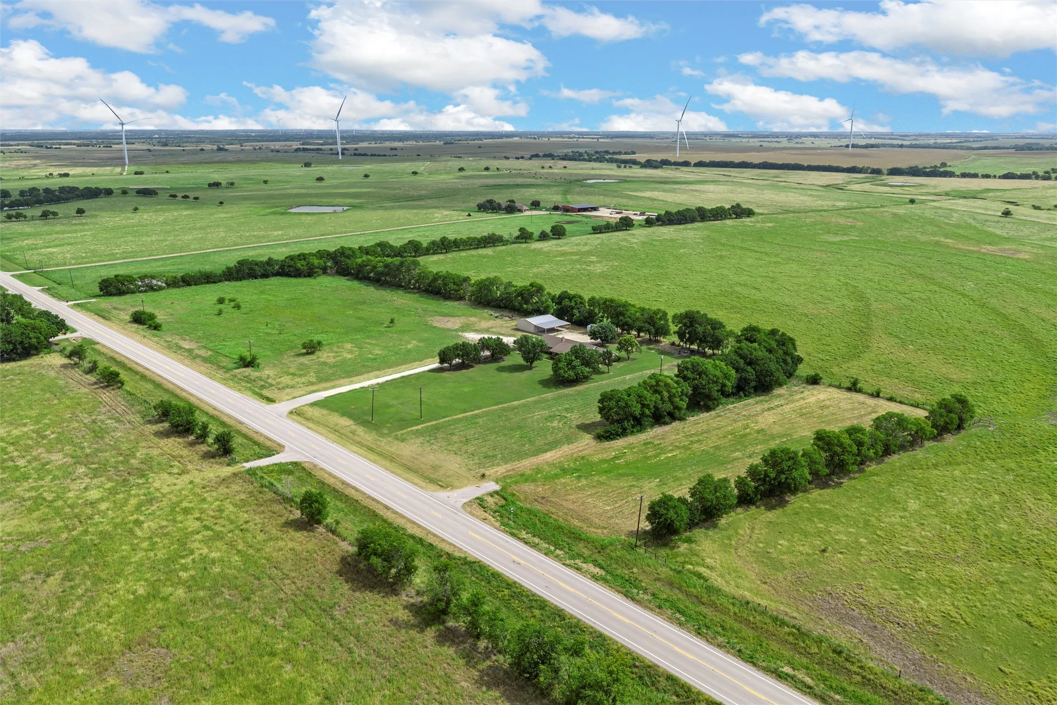 Expansive agricultural land featuring a paved road, scattered trees, and multiple wind turbines on the horizon