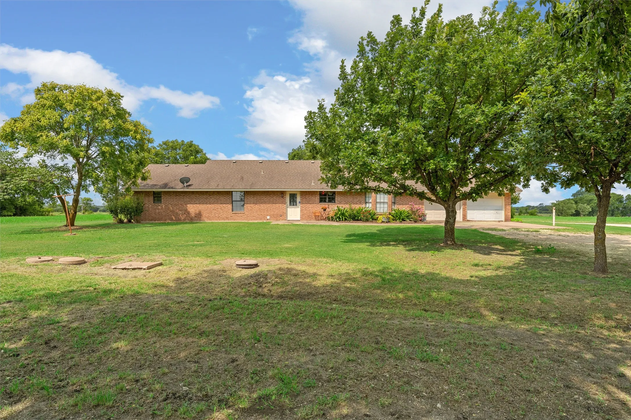 Expansive green lawn surrounds a single-story brick residence with a brown shingled roof, mature trees providing shade, and a double-bay garage