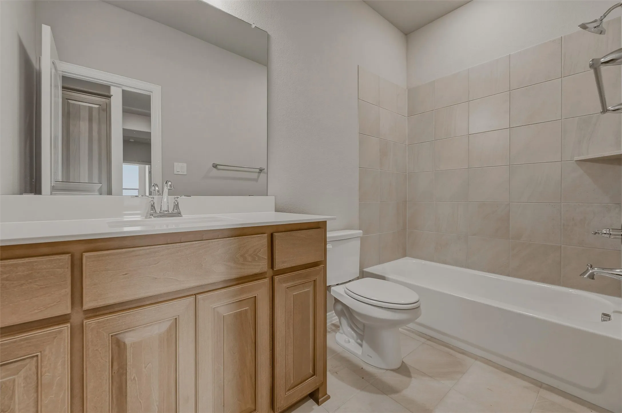 Bathroom featuring a light wood vanity, white countertop, integrated sink with a chrome faucet, full-width mirror, and a tile shower surround