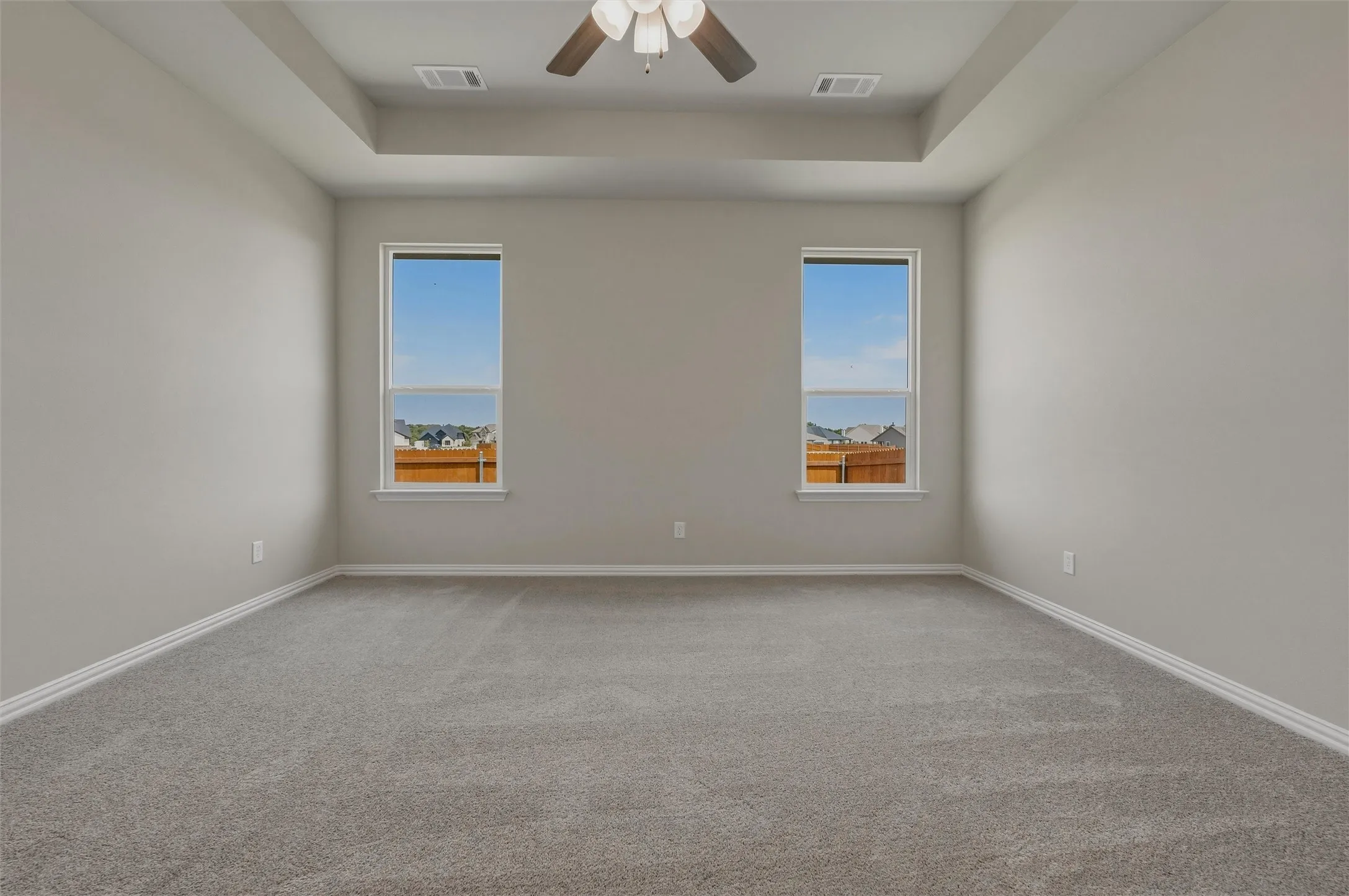 Tray ceiling with an integrated ceiling fan, two windows, and neutral carpeting