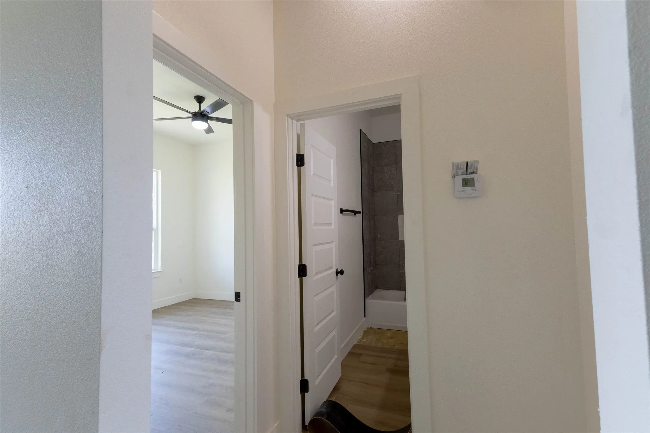 Modern ceiling fan with integrated lighting, wood-finish flooring, white doors with black hardware, and a bathroom featuring a tiled shower surround