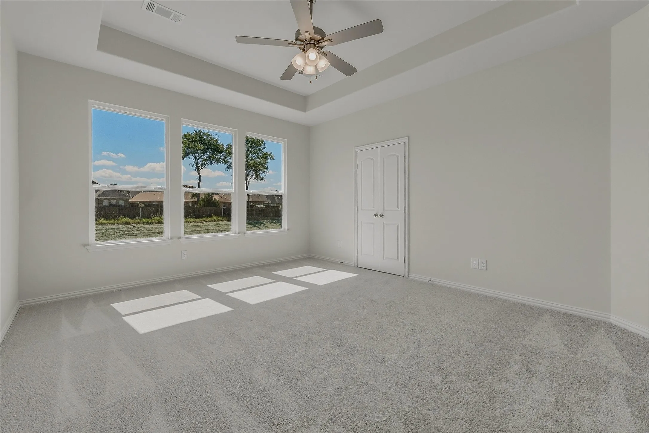 Tray ceiling with a ceiling fan, three expansive windows, light gray carpeting, and a double-door closet