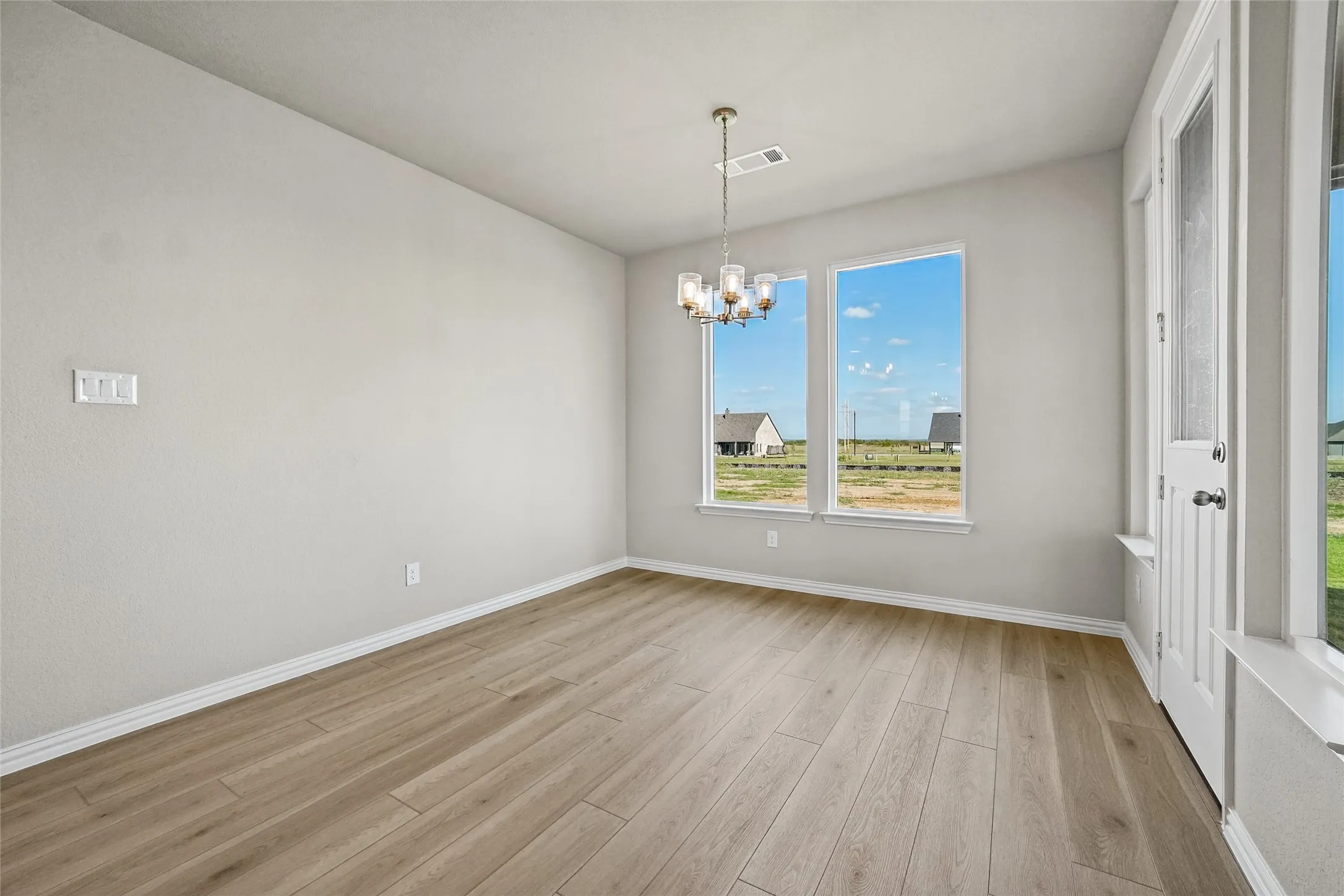Neutral-toned interior featuring wood-finish flooring, dual windows with exterior landscape, a multi-arm pendant light fixture, and a glass-paneled exterior door