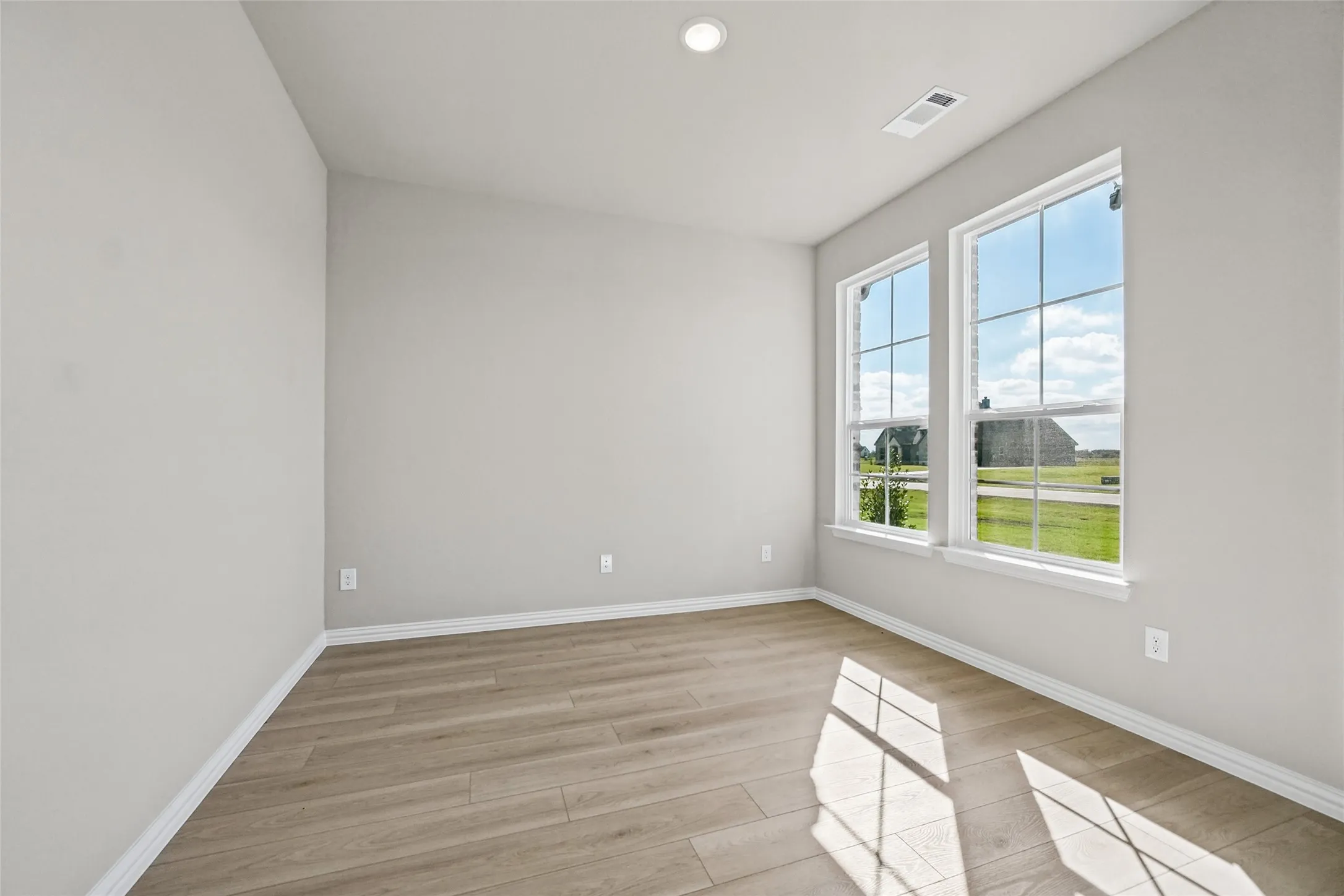 Bright, naturally lit bedroom showcasing wood-finish flooring, recessed ceiling lighting, and large double-pane windows framing pastoral scenery
