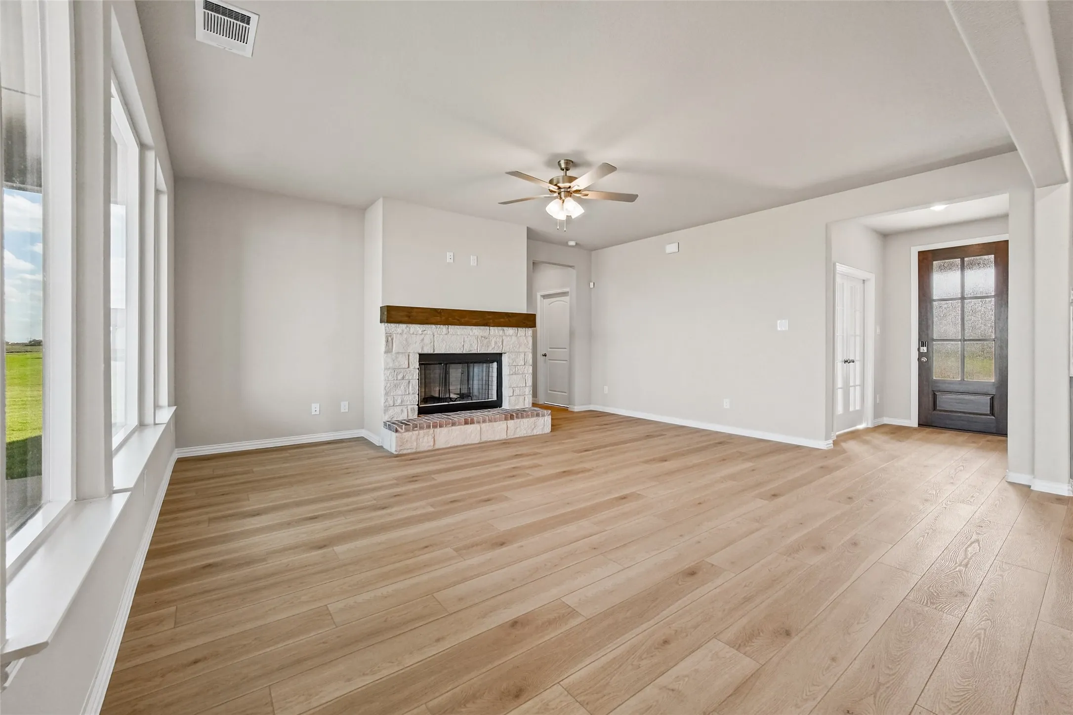 Living area featuring wood-finish flooring, a stone-surround fireplace with a timber mantel, a multi-pane glass entry door, and large windows