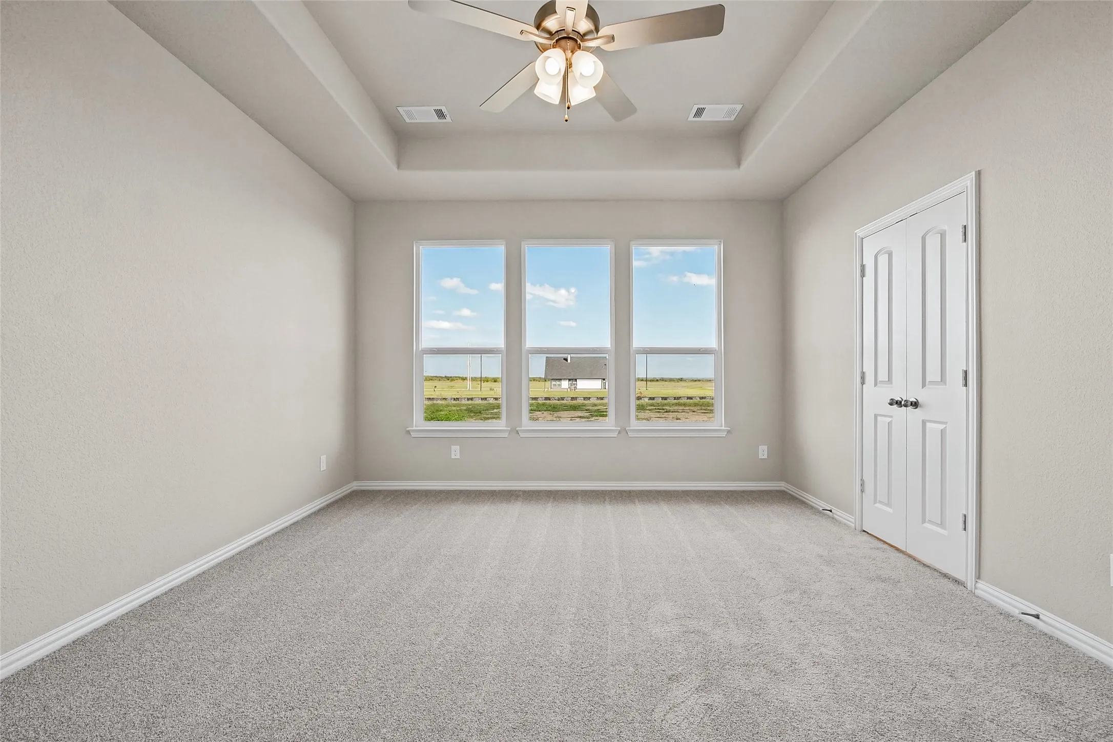 Tray ceiling with a central multi-light ceiling fan, neutral carpeting, and a trio of windows providing natural light through double-pane glass