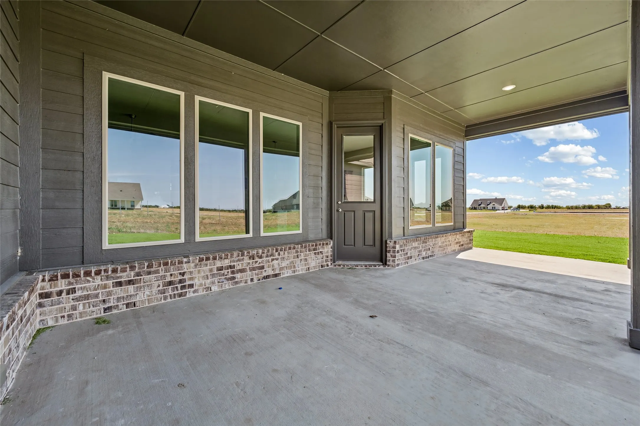 Expansive covered porch with polished concrete flooring, brick veneer wainscoting, and multiple large multi-pane windows framing open countryside