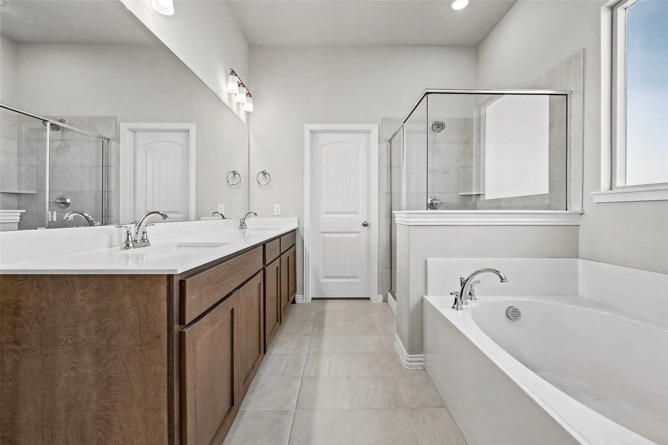 Dual vanity with wood-finish cabinetry and white countertops, walk-in shower with glass enclosure, white soaking tub, and neutral tiled flooring