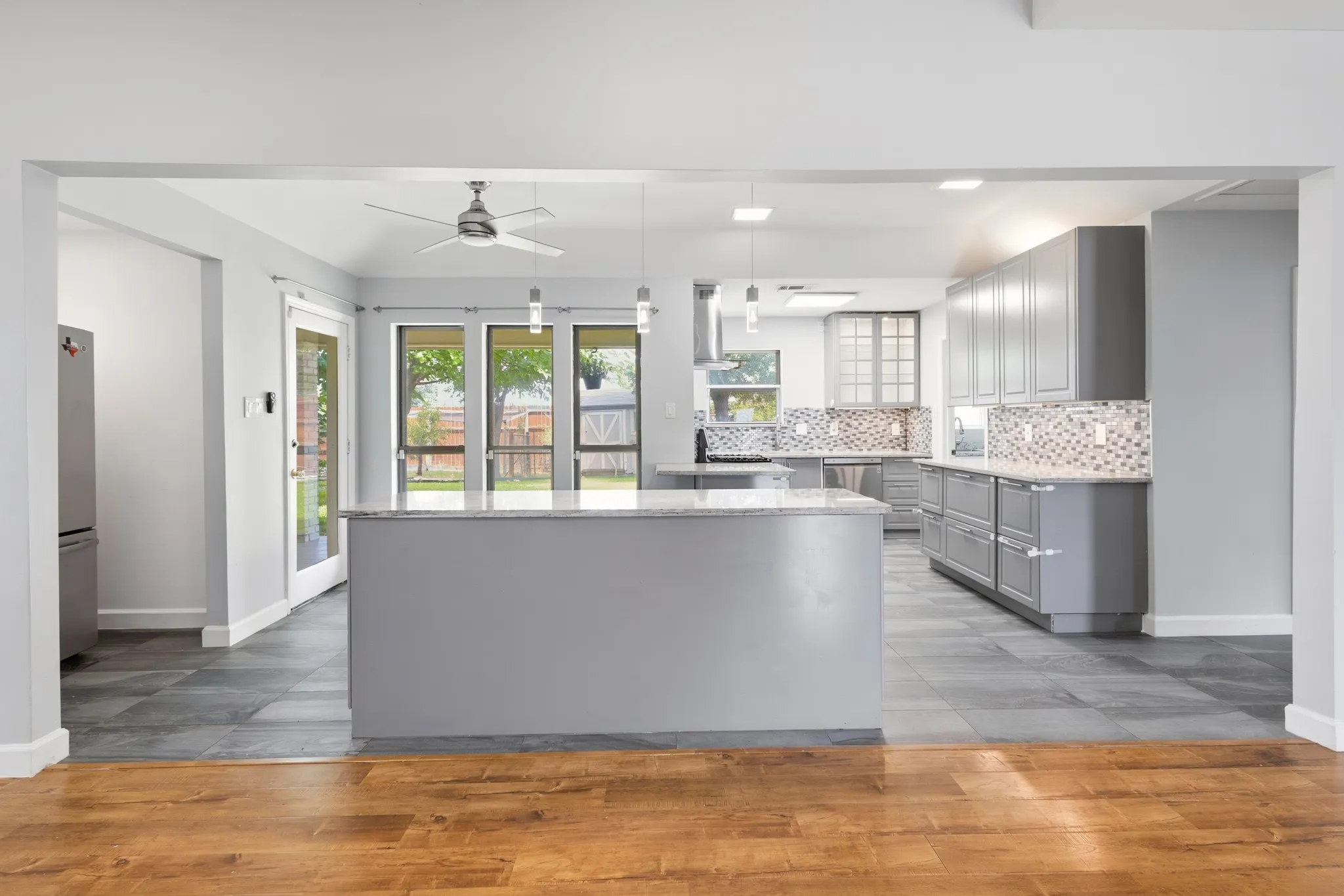 Spacious kitchen featuring an expansive center island with a light-toned countertop, gray cabinetry, a decorative tile backsplash, recessed lighting, and a stainless steel refrigerator