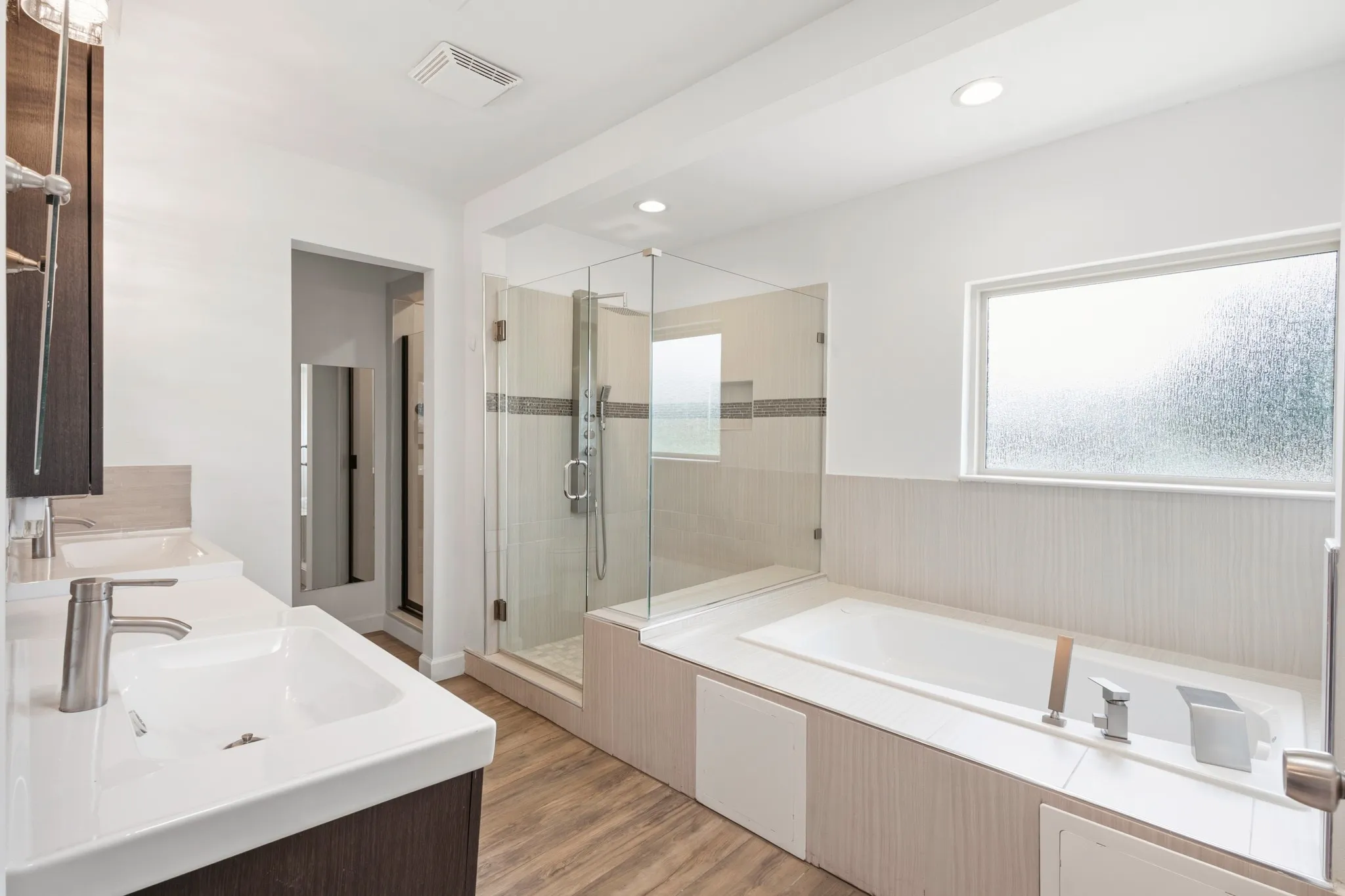 Contemporary bathroom featuring a spacious walk-in shower with glass enclosure, a built-in soaking tub, dual vanities with integrated sinks, wood-finish flooring, and a privacy window
