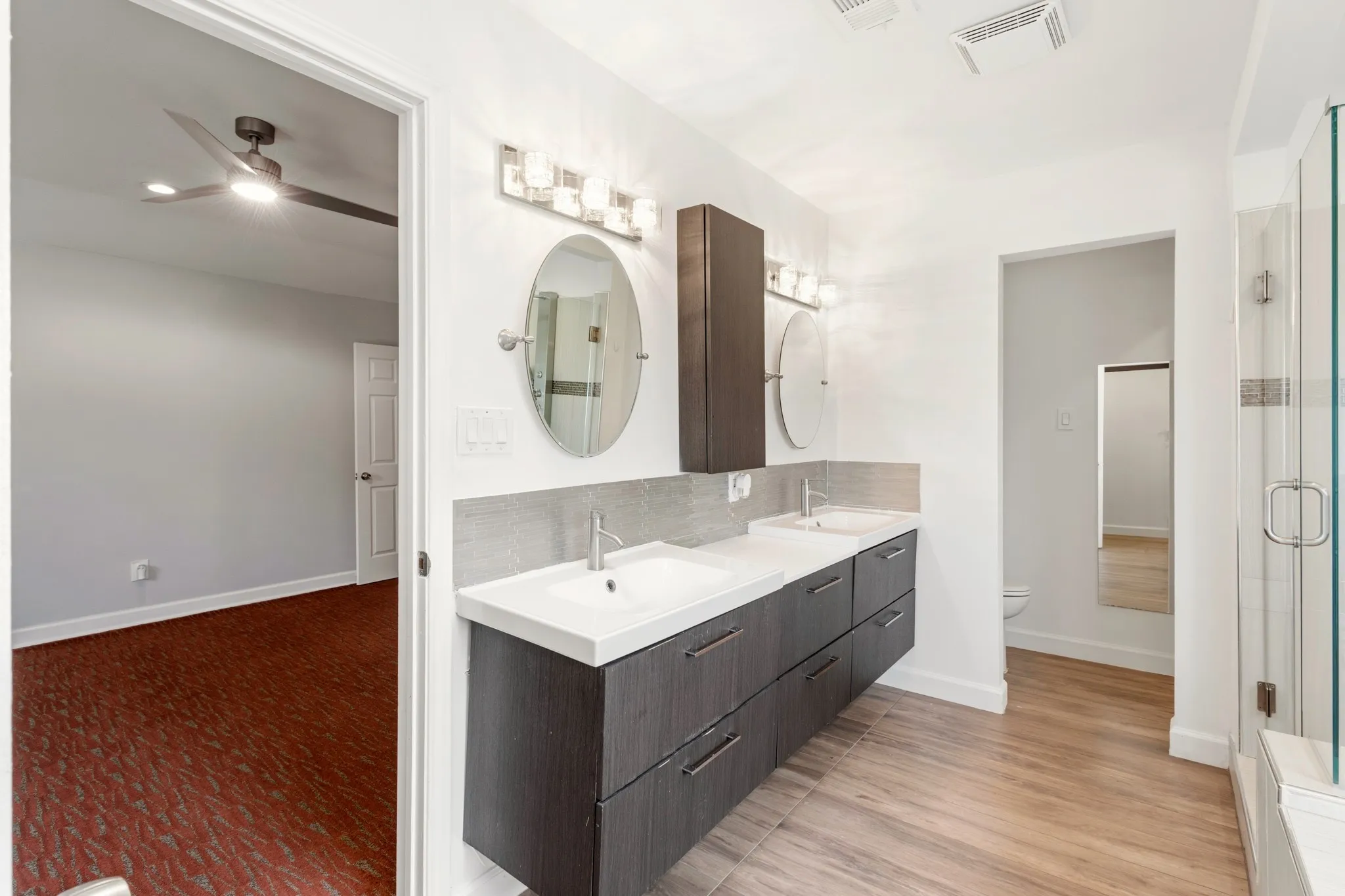 Dual vanity with white countertops and undermount sinks, wood-finish flooring, glass-enclosed shower, two oval mirrors, and contemporary lighting fixtures