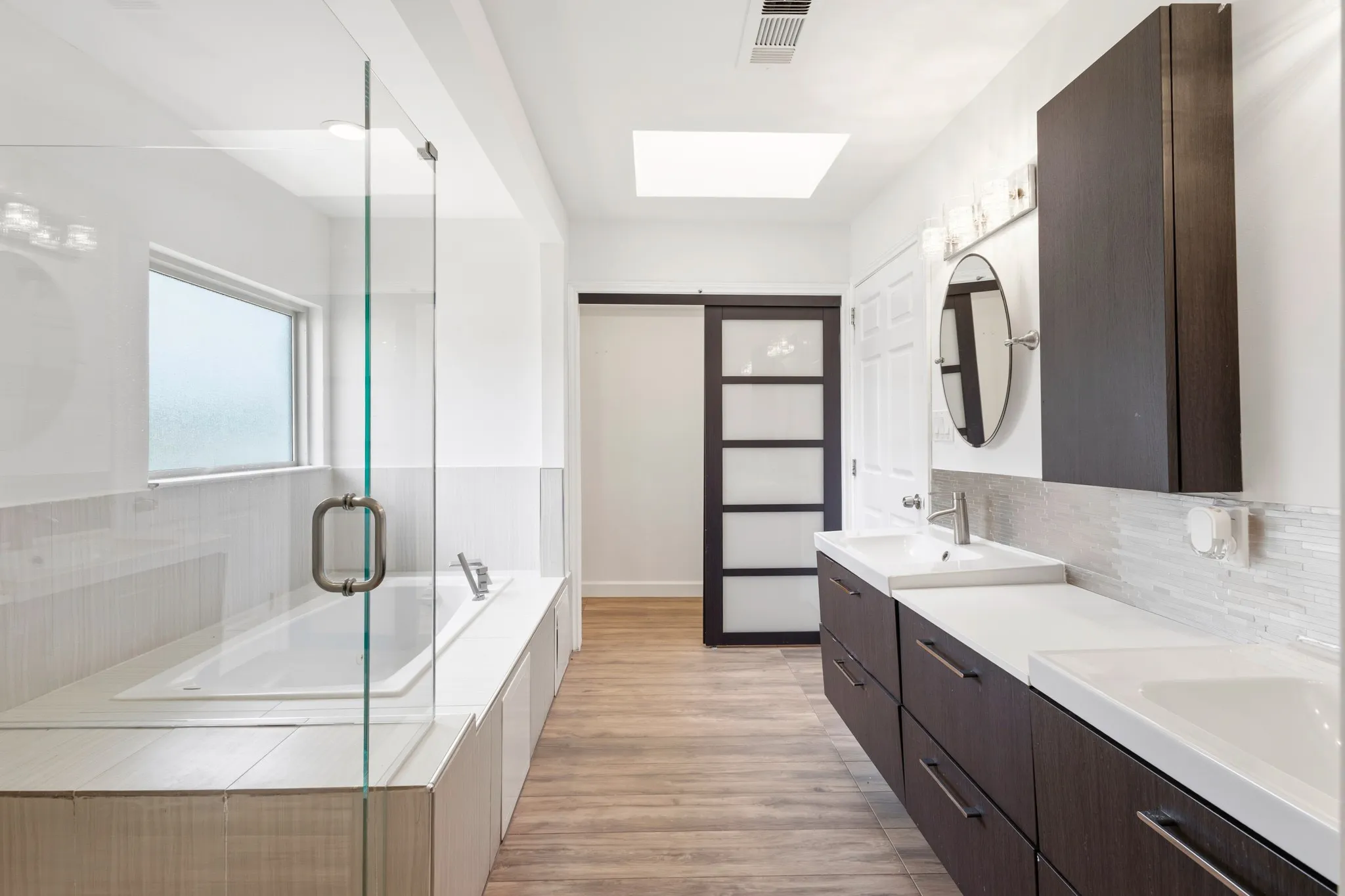 Contemporary bathroom featuring a spacious built-in tub, a glass-enclosed shower, a double vanity with integrated sinks, wood-finish flooring, and a skylight