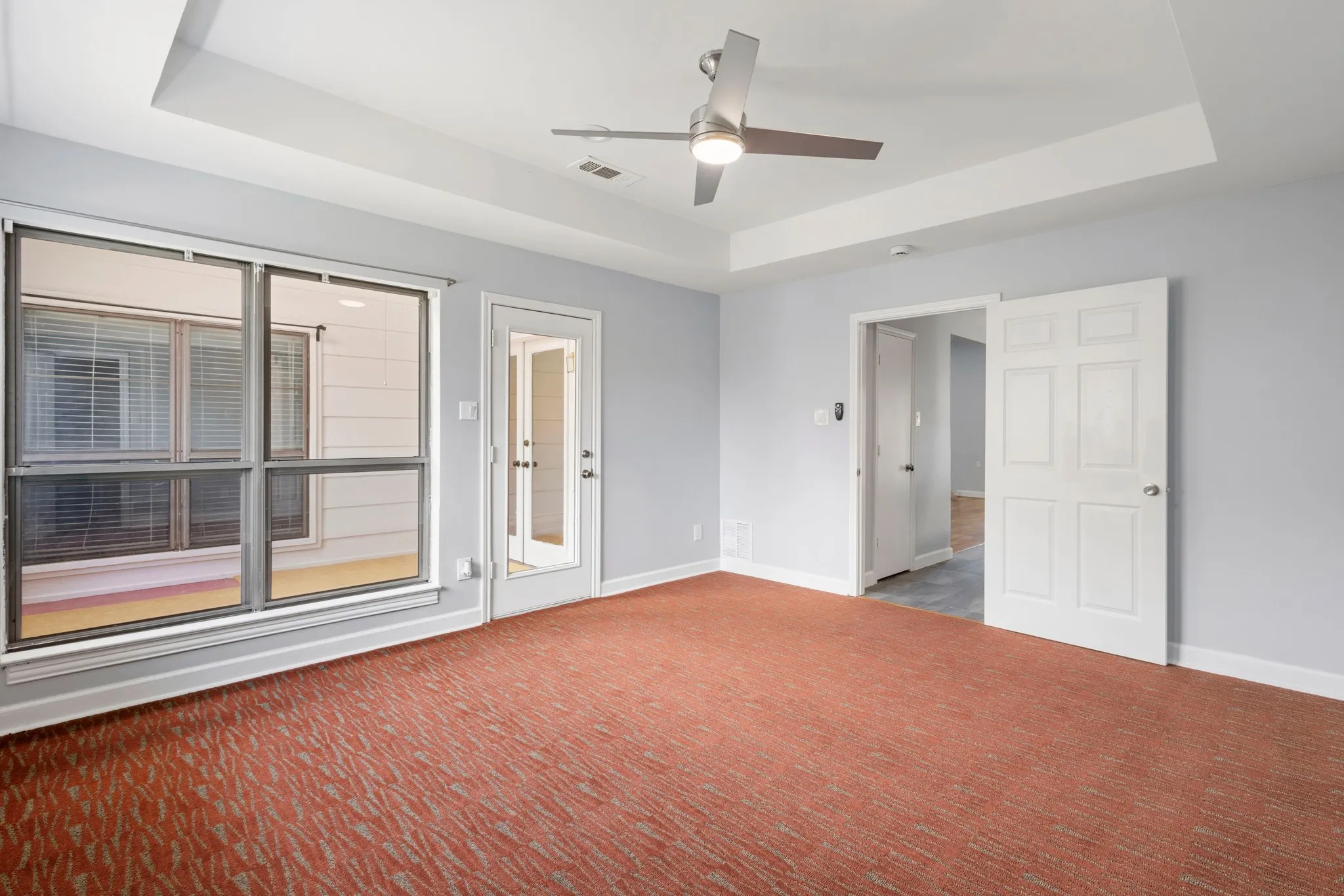 Spacious bedroom room featuring a large window with blinds, recessed ceiling lighting, a paneled interior door, light blue wall paint, and vibrant patterned carpeting