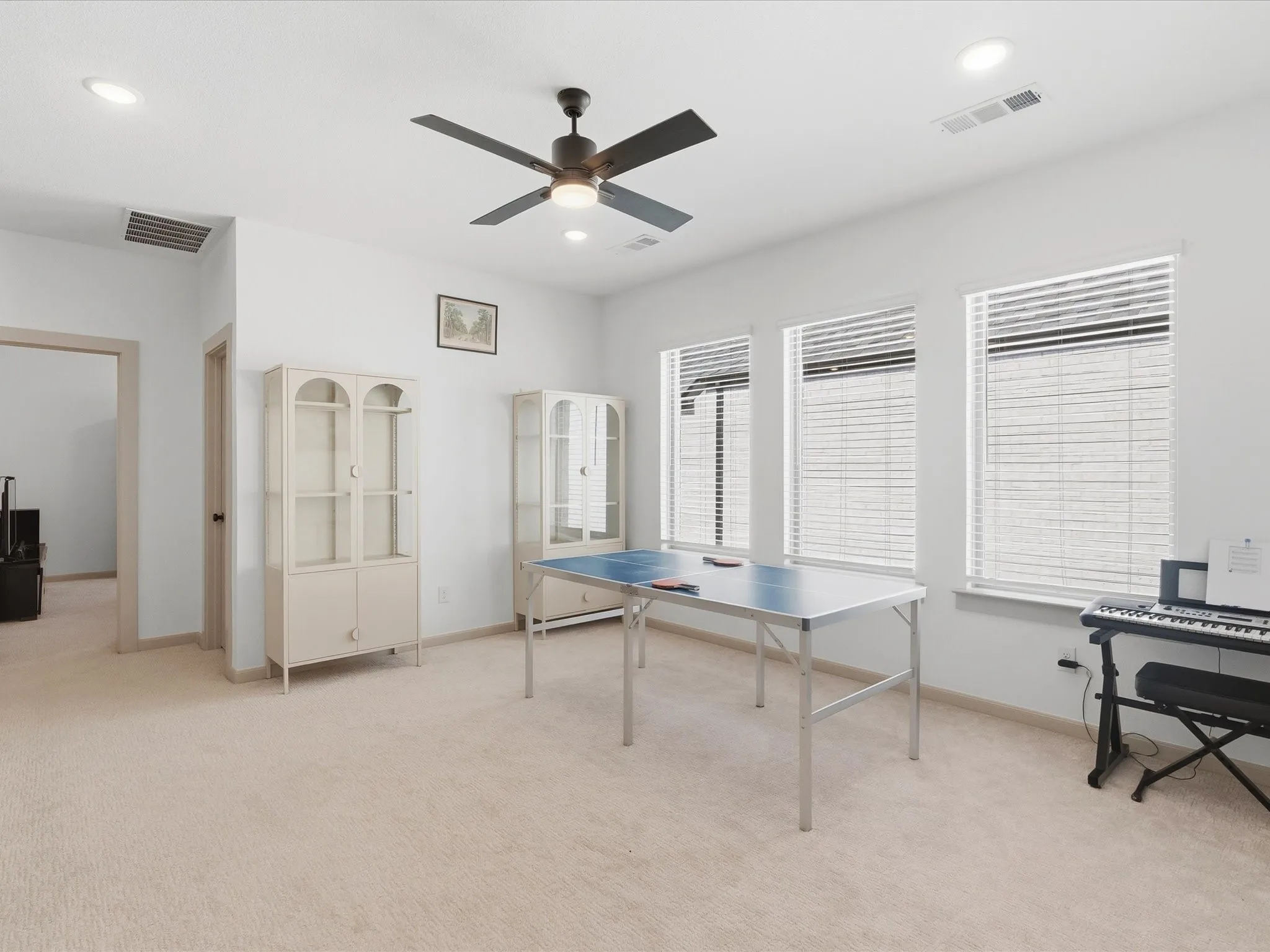 Spacious room featuring light-colored carpeting, multiple windows with blinds, a ceiling fan with integrated lighting, and recessed ceiling lights