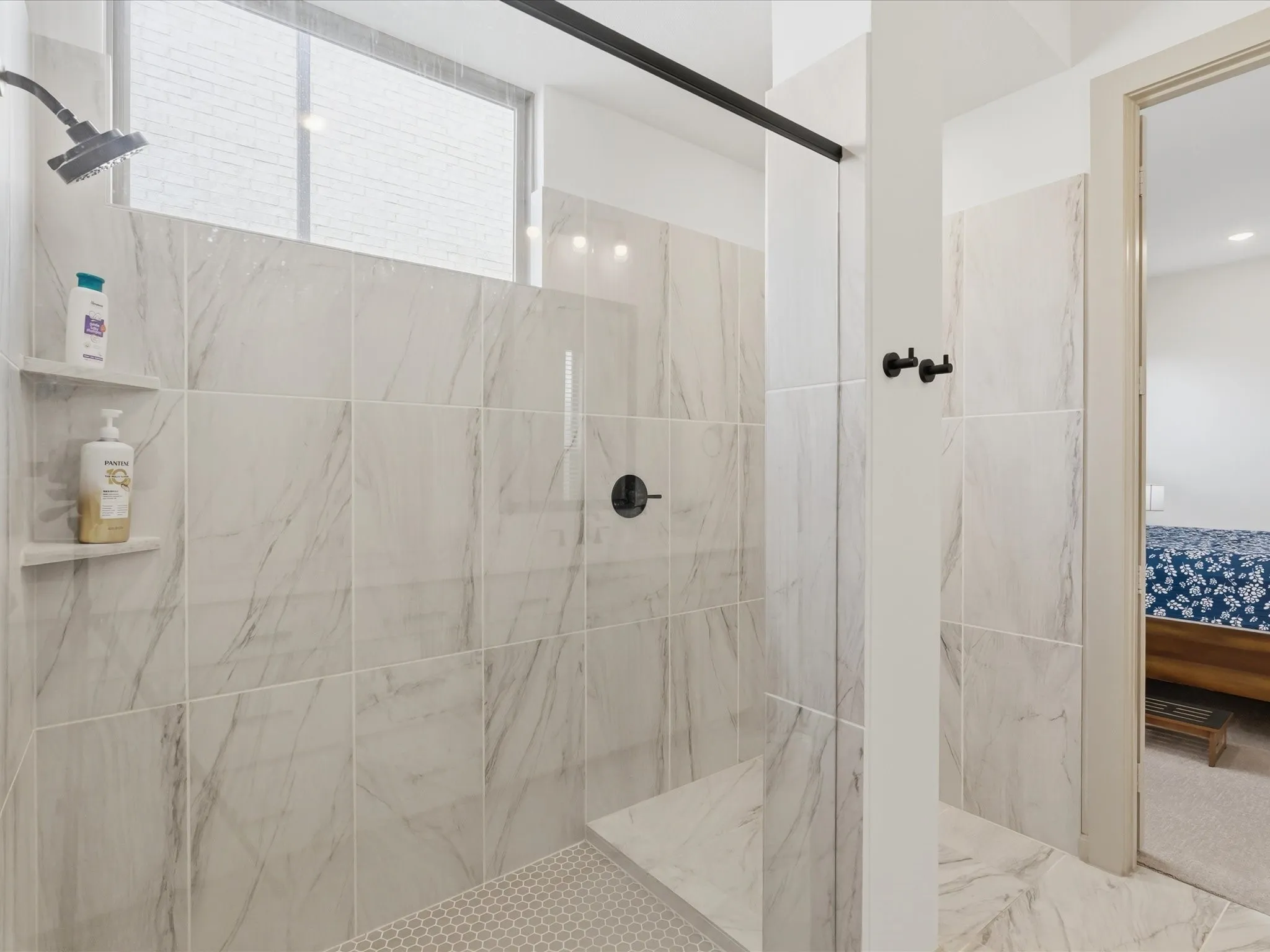 Walk-in shower featuring large format marbled tile, built-in shelving, a shower bench, hexagonal mosaic floor tile, and a black-finish shower head and controls