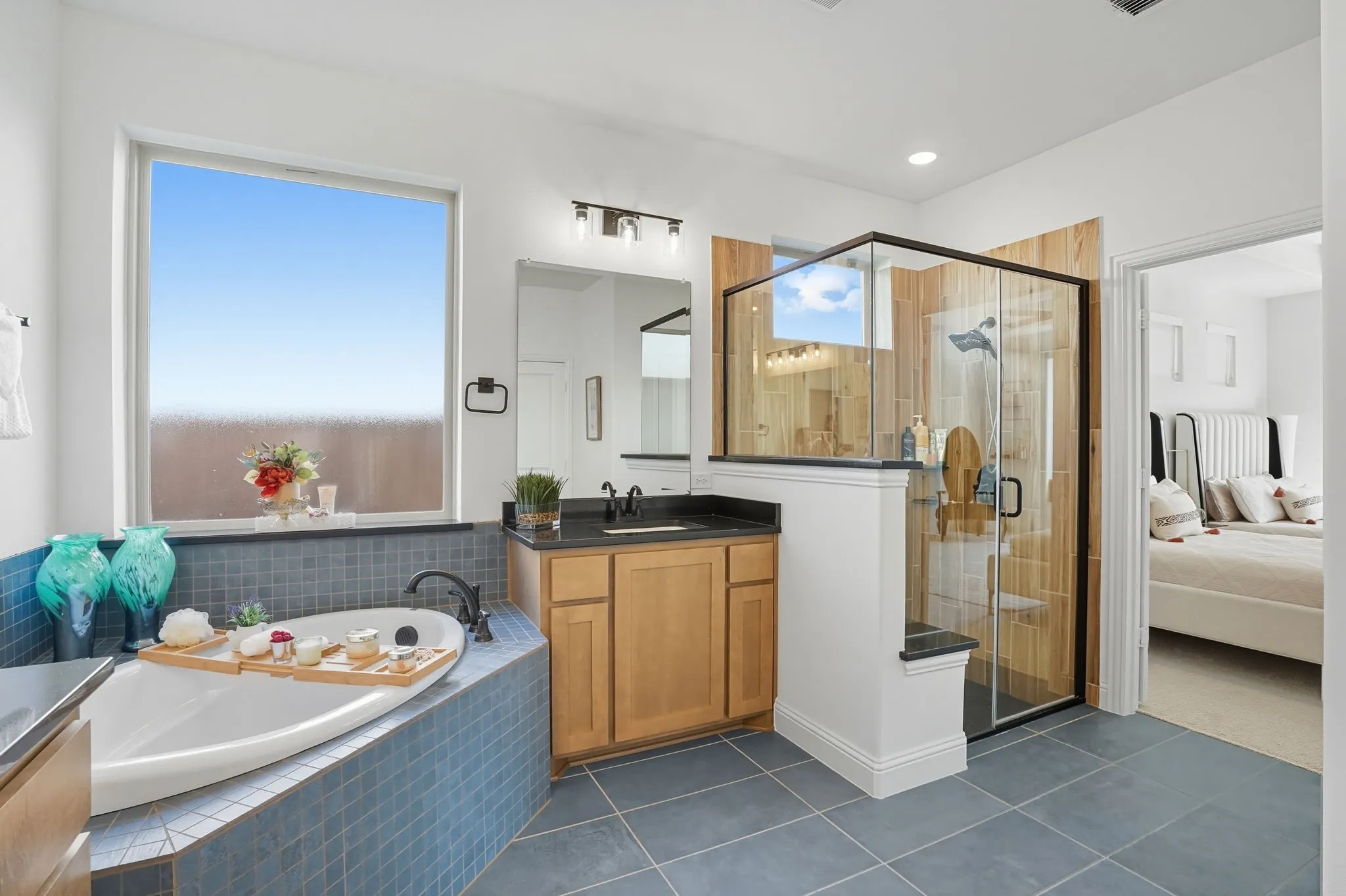 Spacious bathroom featuring a built-in soaking tub with blue tile surround, a glass-enclosed shower with wood-finish wall panels, a single vanity with wood cabinetry and dark countertop, and large format floor tiling