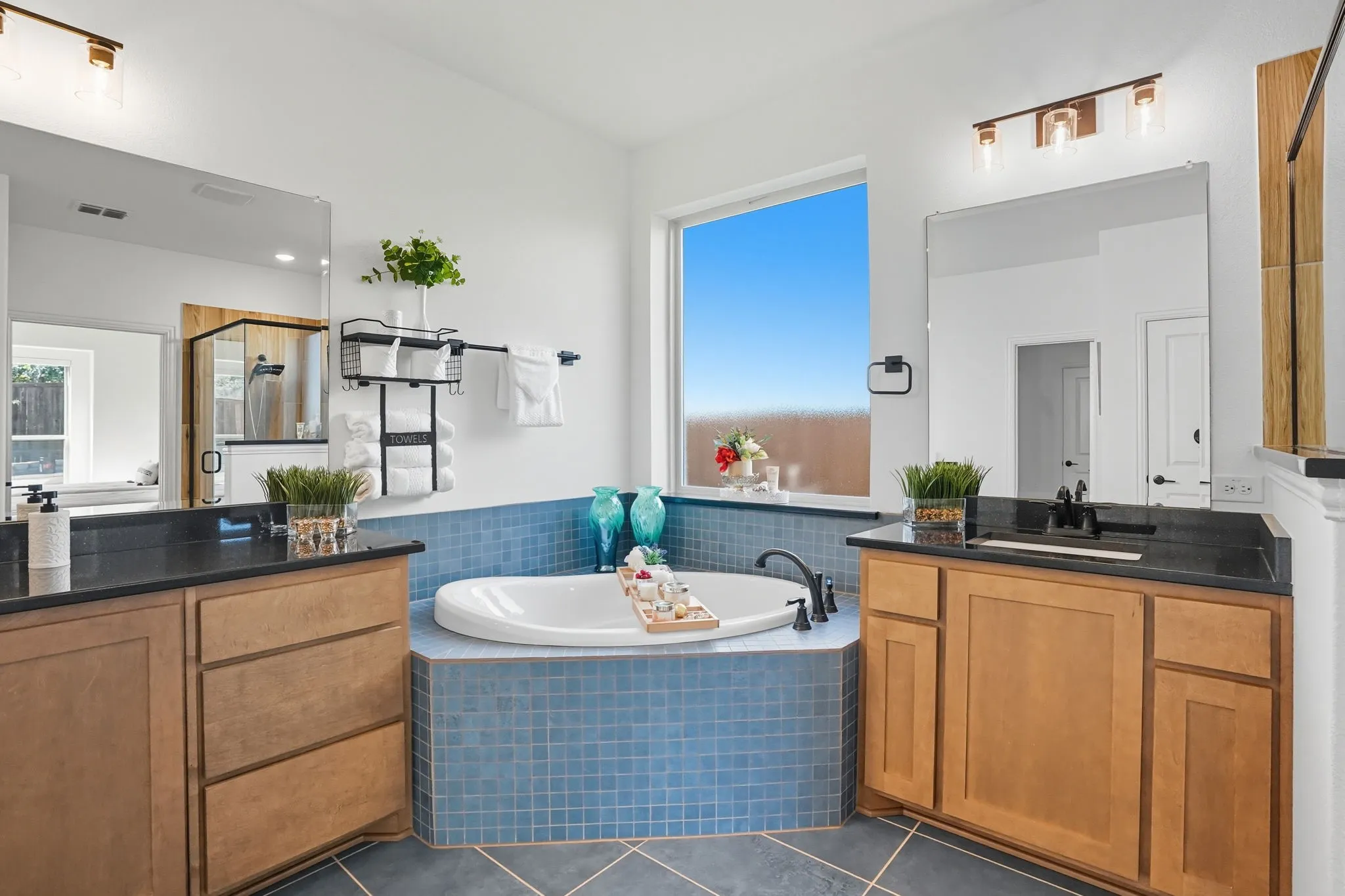 Corner soaking tub with blue tile surround, flanked by twin wood-finish vanities with dark countertops