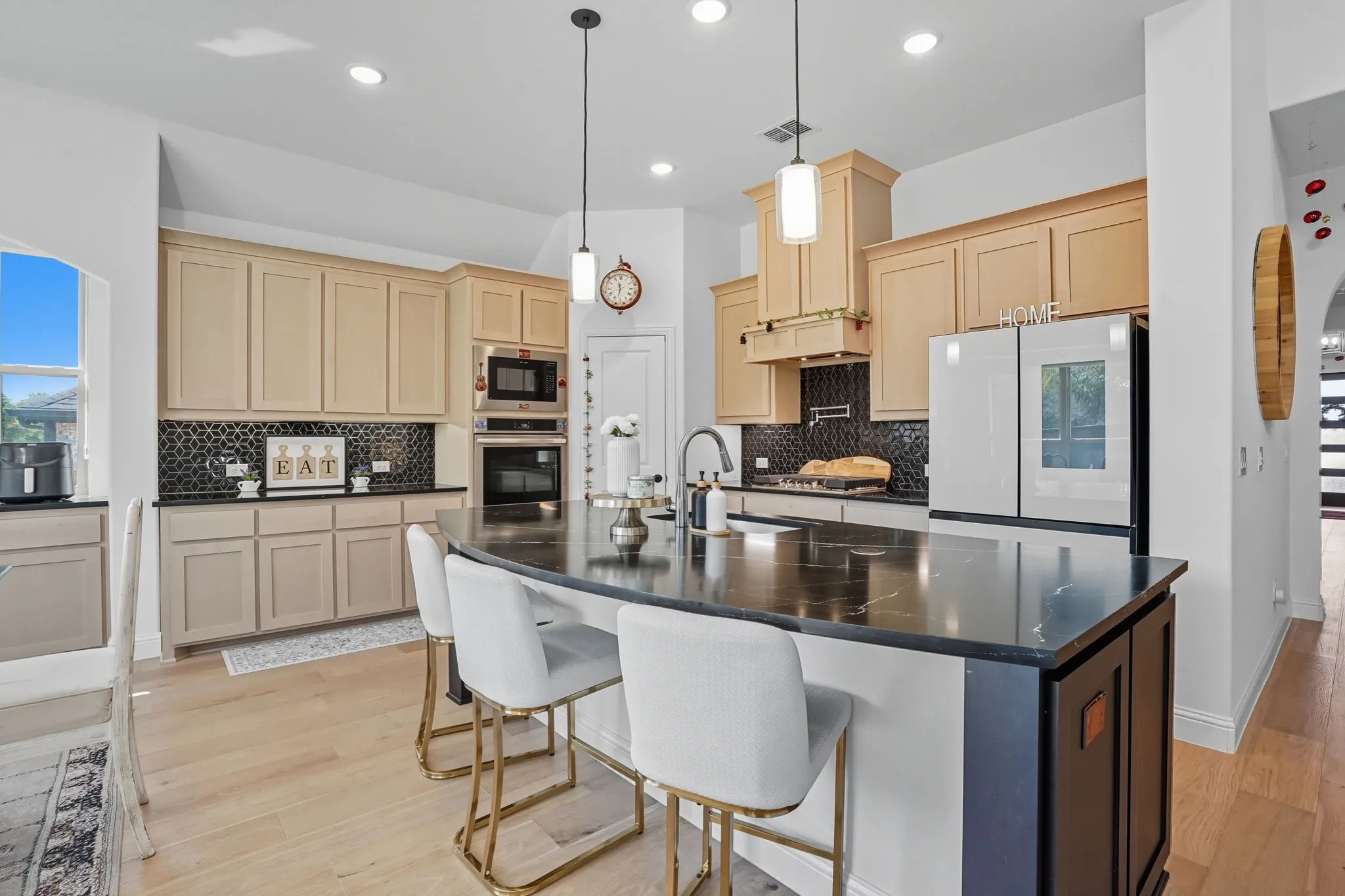 Spacious kitchen featuring a large island with seating, light wood-finish flooring, built-in stainless steel appliances, neutral cabinetry, and a dark mosaic tile backsplash