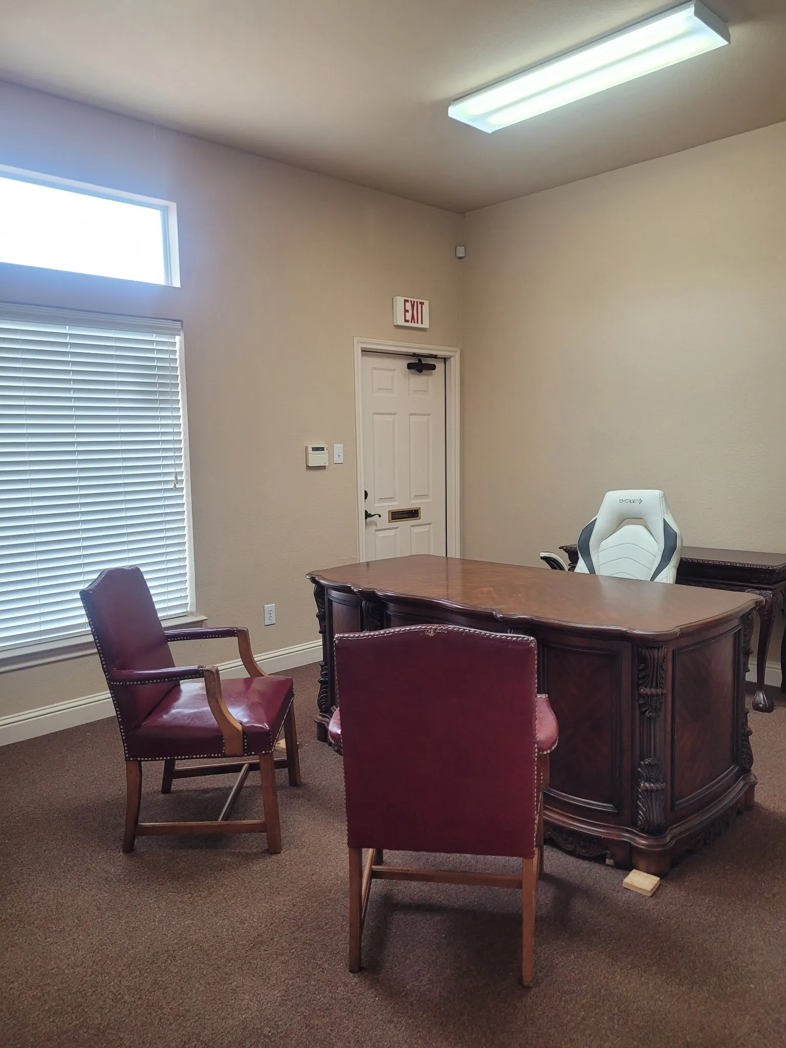 Spacious room featuring a decorative wood-finish desk, neutral wall paint, and a large window with horizontal blinds