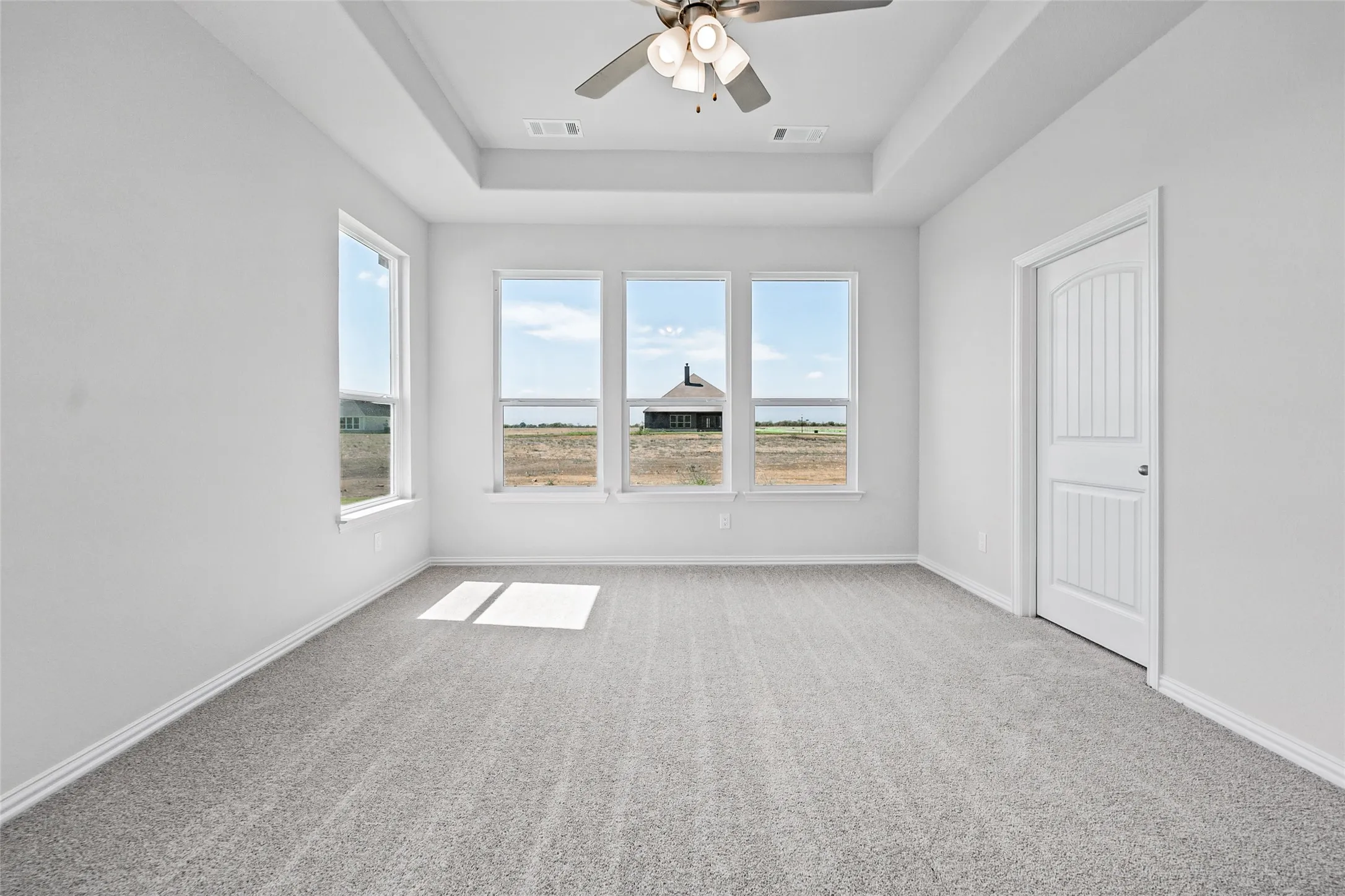 Recessed tray ceiling, neutral tone carpeting, three large windows, and a ceiling fan with integrated lighting