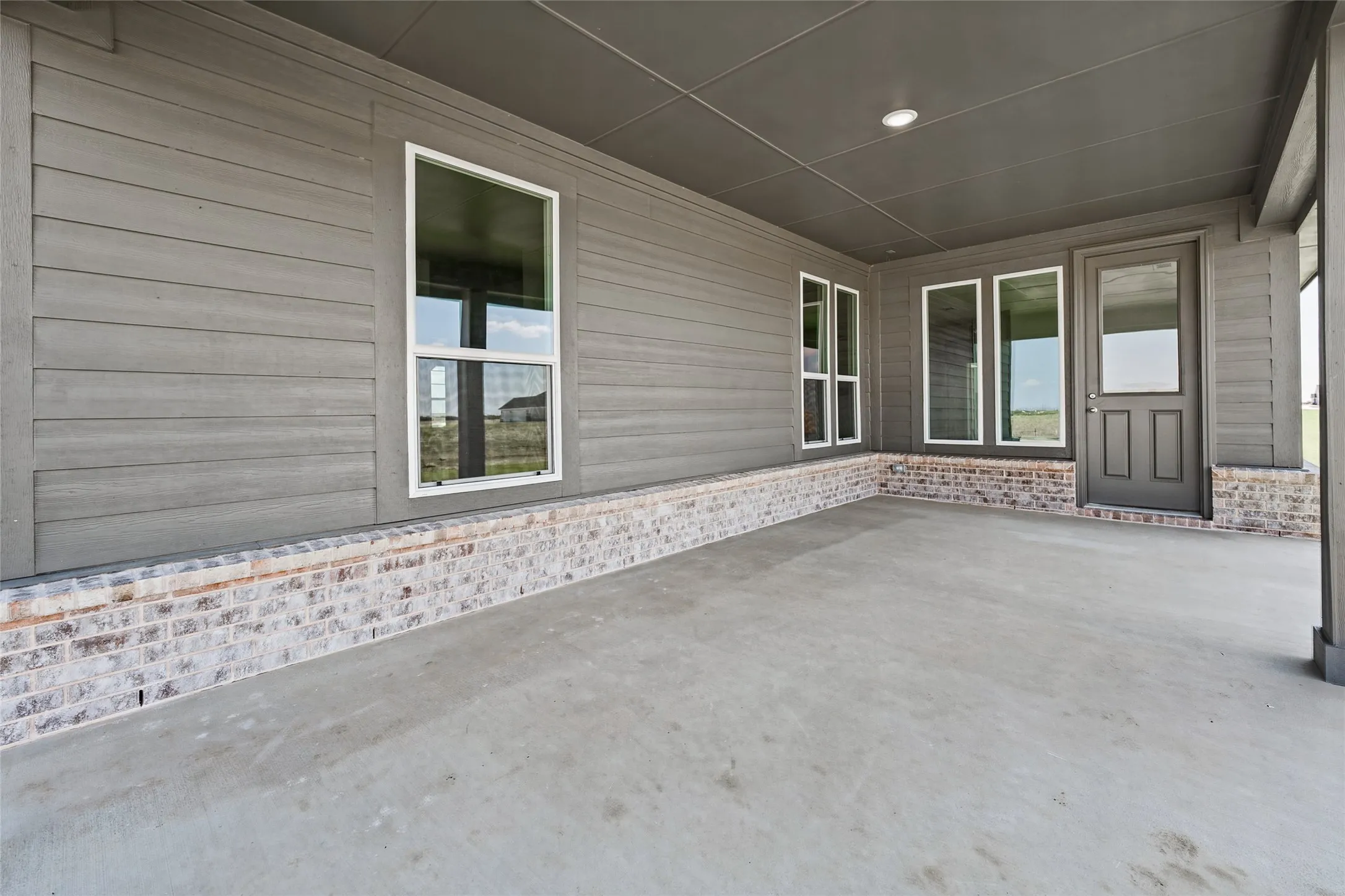Covered concrete patio featuring shiplap siding, a brick perimeter, and multiple windows