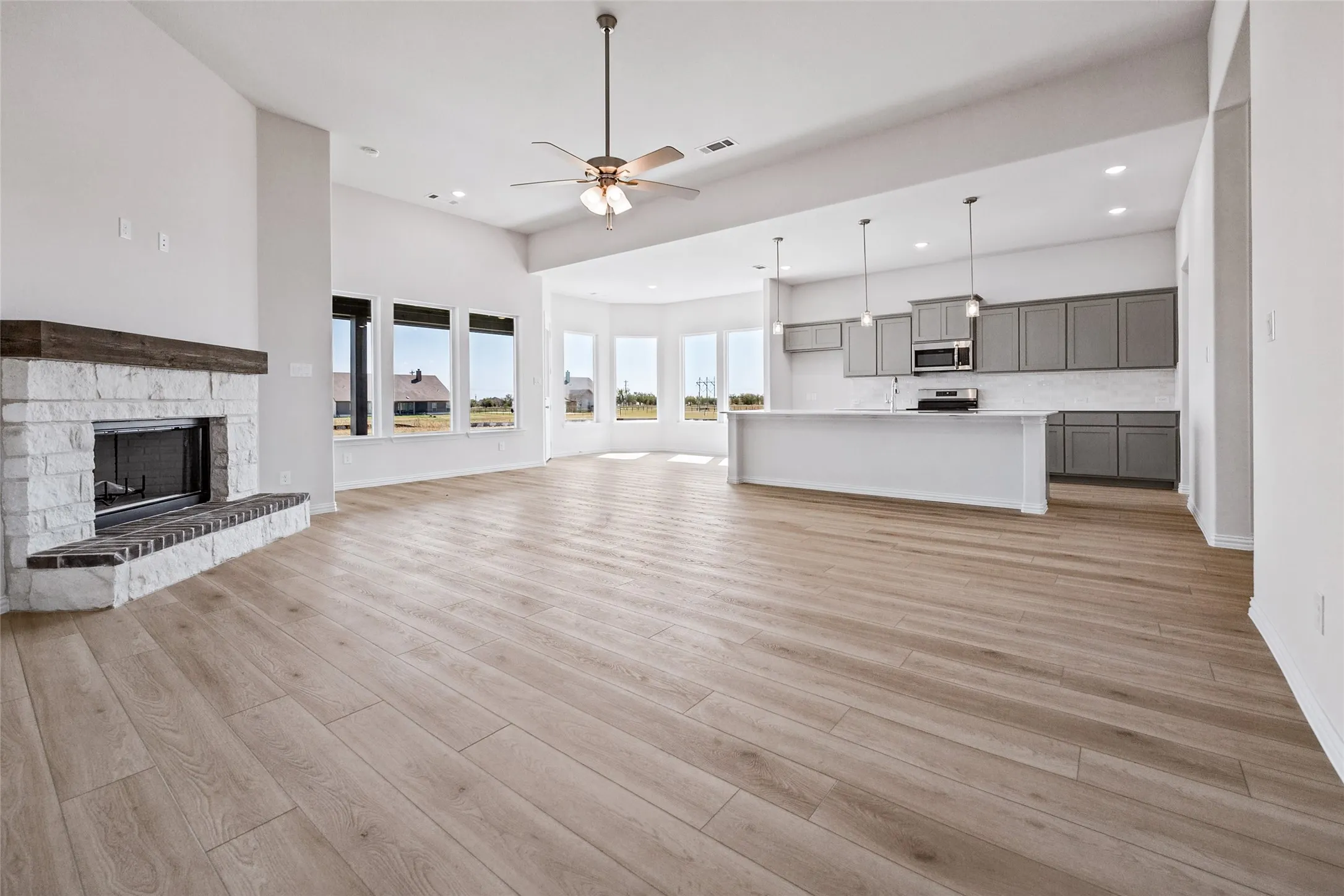 Open-concept living area featuring wood-finish flooring, a stone fireplace with a wood mantle, and an expansive kitchen island with pendant lighting