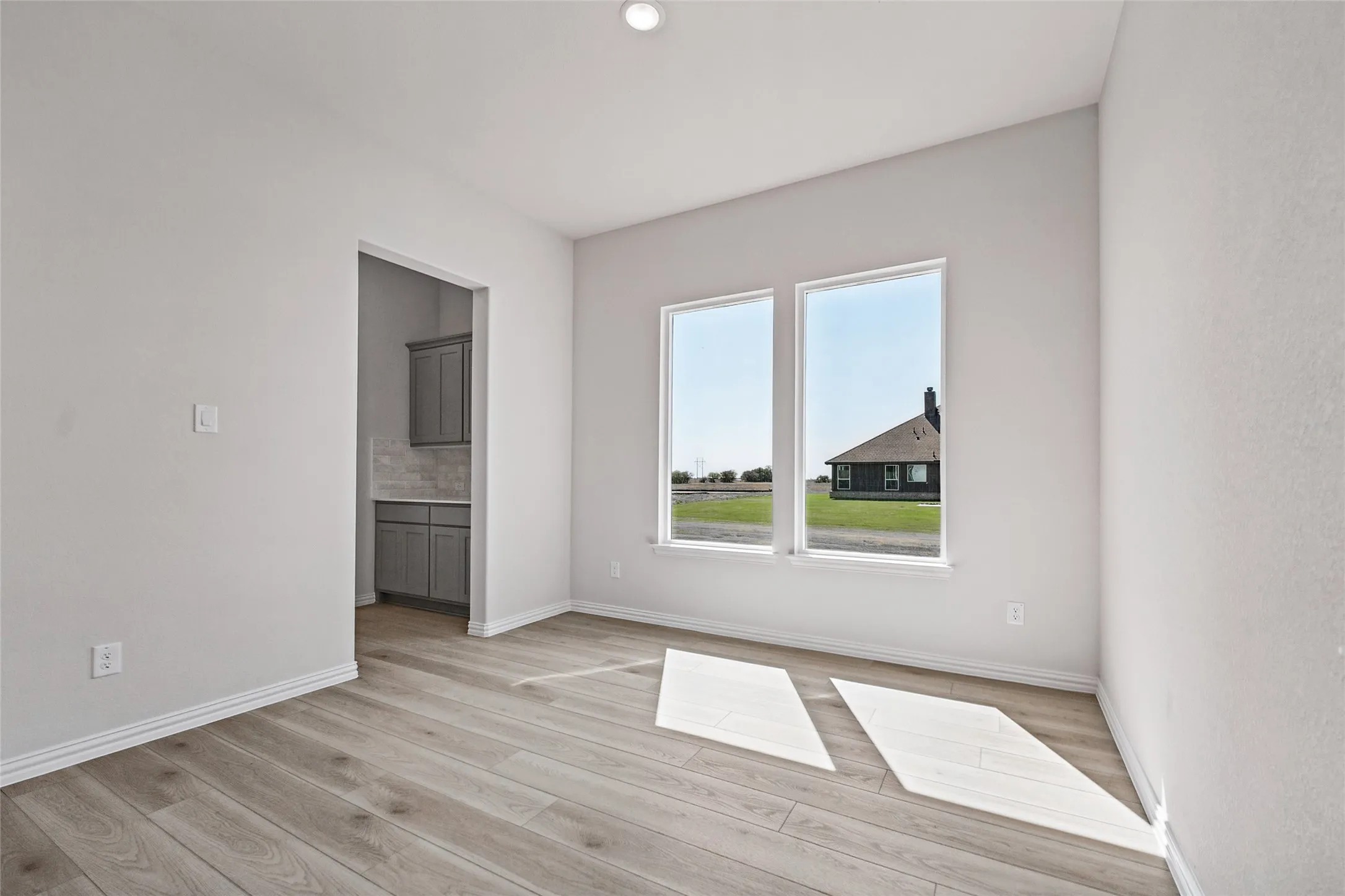 Recessed ceiling lighting, wood-finish flooring, two large windows, white walls, and a separate space with dark gray cabinetry and a tiled backsplash