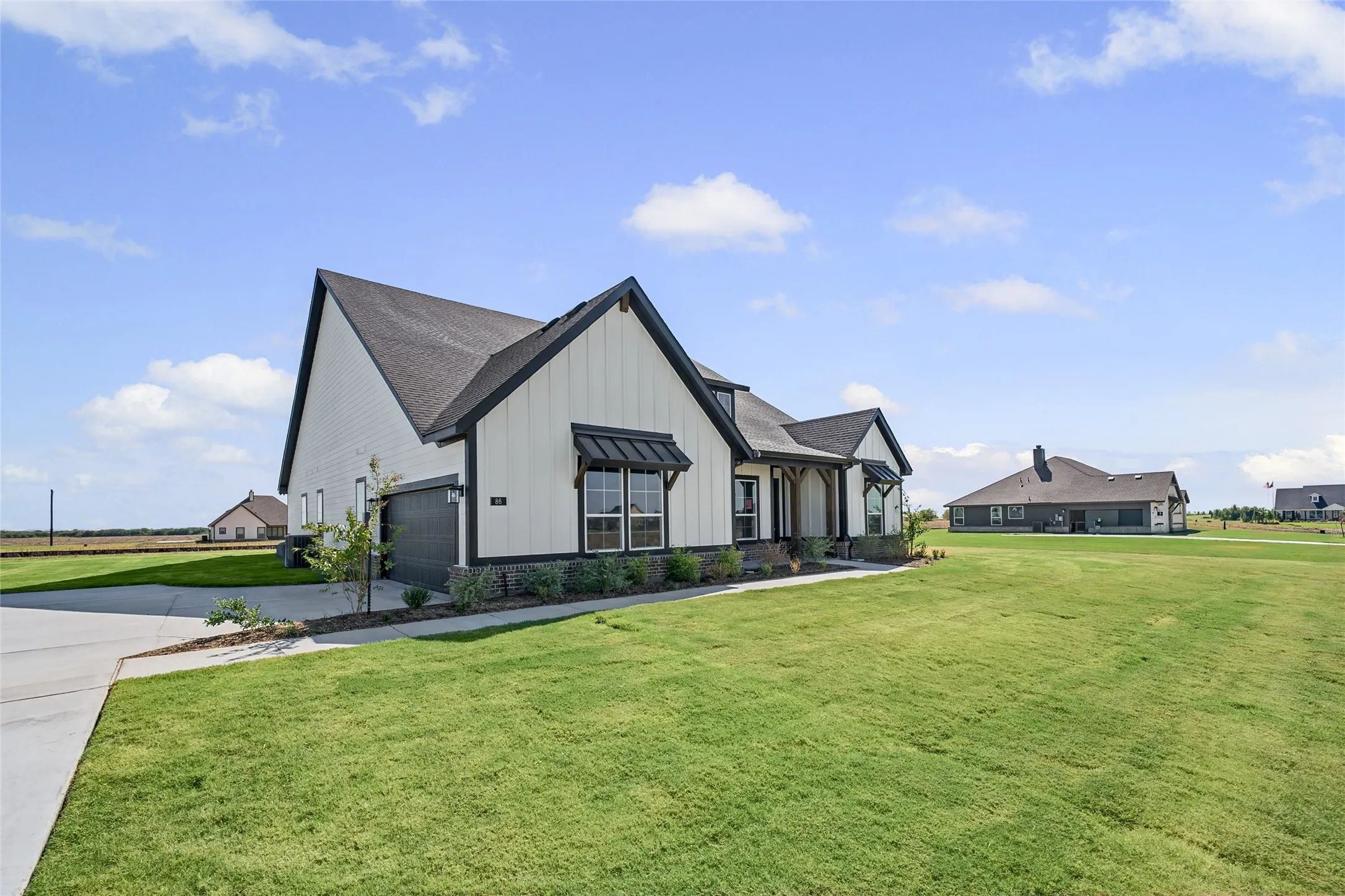 Modern farmhouse estate featuring a striking black and white exterior with peaked rooflines, expansive manicured lawn, concrete driveway, and multiple outbuildings positioned on sprawling acreage with clear sightlines across the property