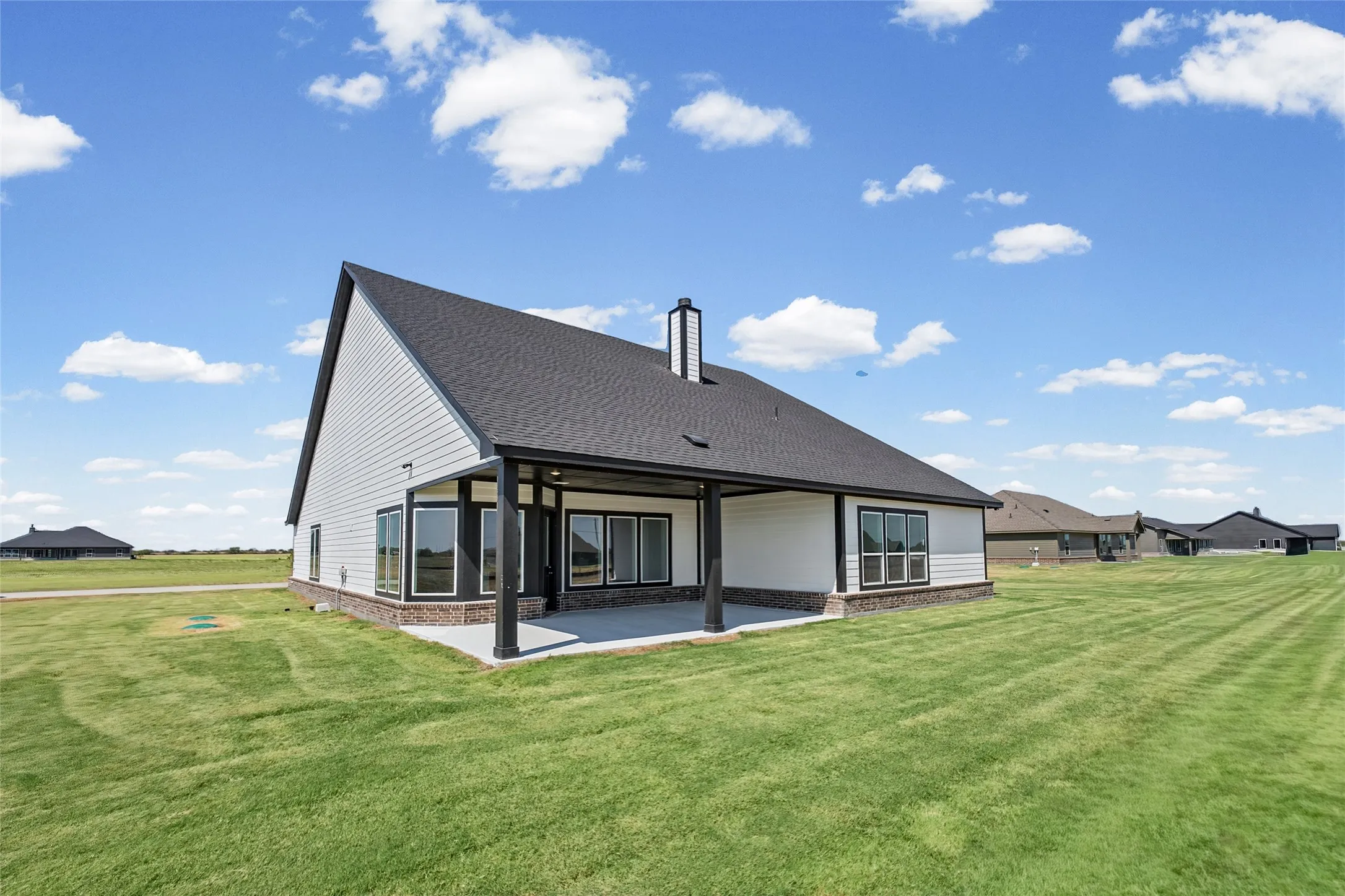 Expansive lawn with a covered patio, dark shingle roof, white siding, and brick veneer detailing