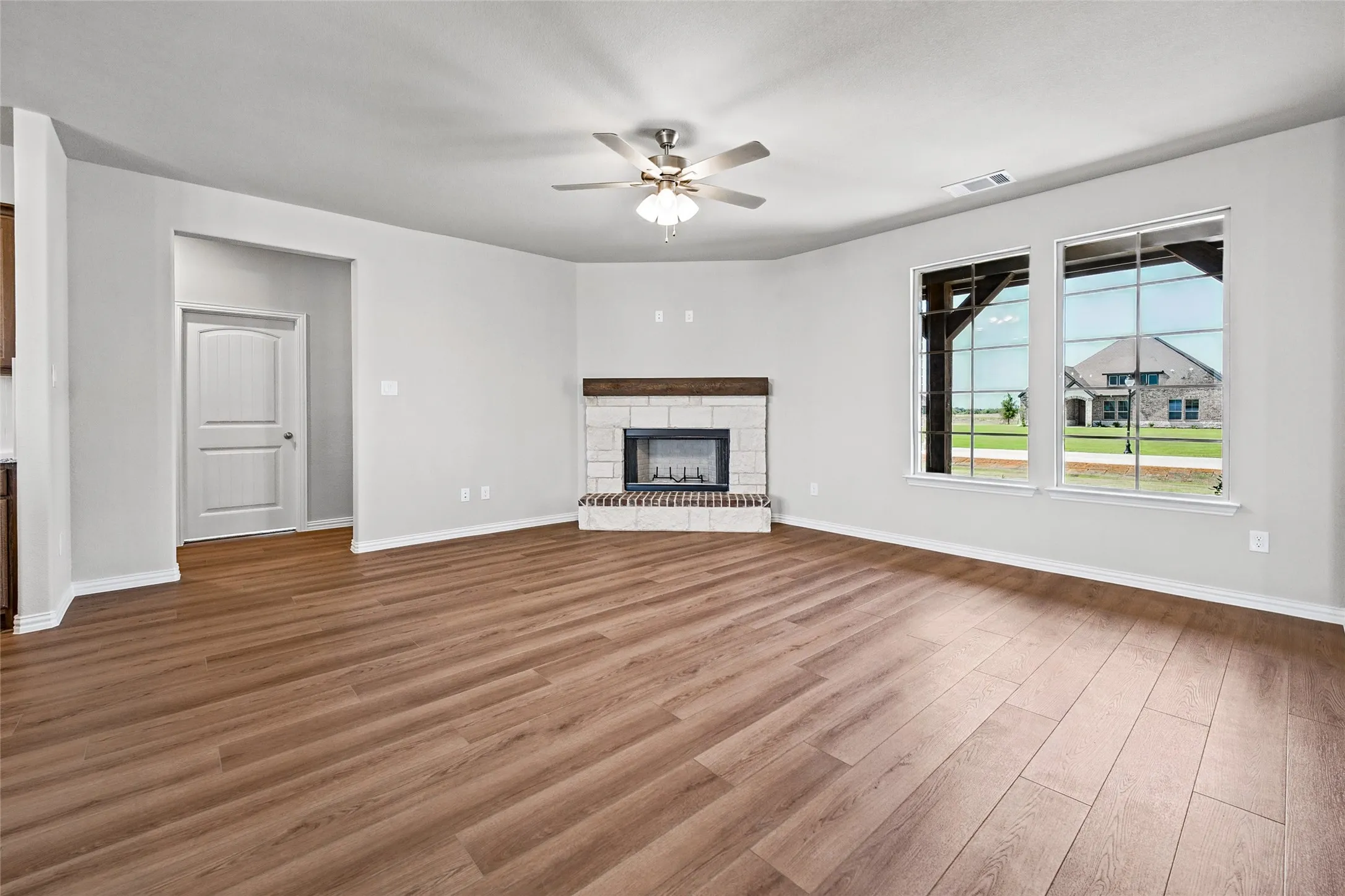 Spacious living area featuring wood-finish flooring, a white stone fireplace with a wood mantle, a ceiling fan, and large windows