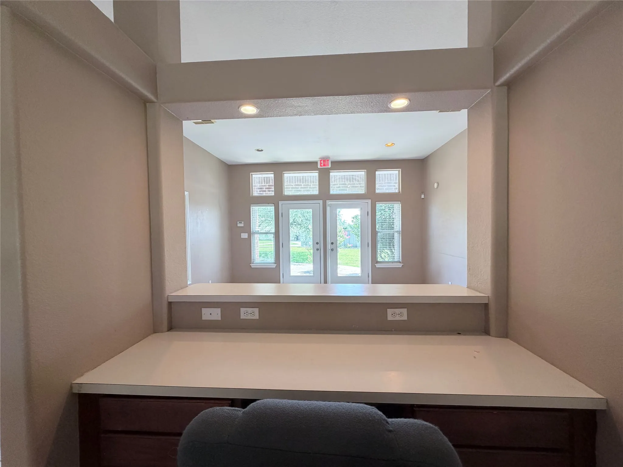 Built-in workspace with a light-toned countertop and dark wood-finish cabinetry, featuring a pass-through opening to a room with recessed lighting and double glass panel doors